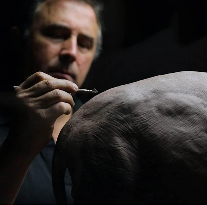 A man working on a large sculpture or model, focusing on the elephant's back as he uses a small tool for detailing.