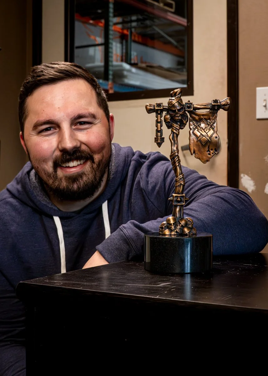 A smiling man with a beard and short hair wearing a dark hoodie, resting his arm on a table, with a Mak'gora dueling flag bronze sculpture from World of Warcraft.
