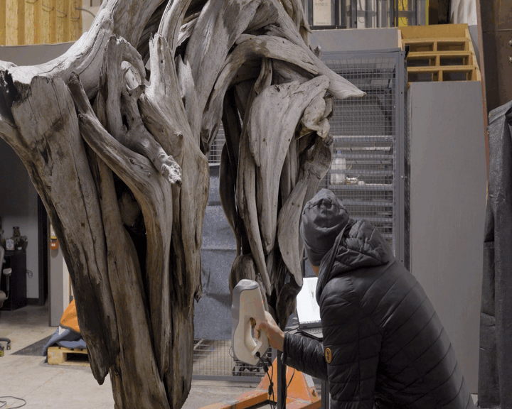 Person in a black jacket and gray beanie taking 3D scans of a large wooden sculpture.