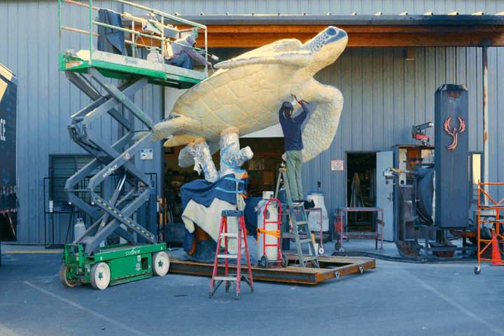People working on a large turtle sculpture outside a building, with equipment and scaffolding around them.