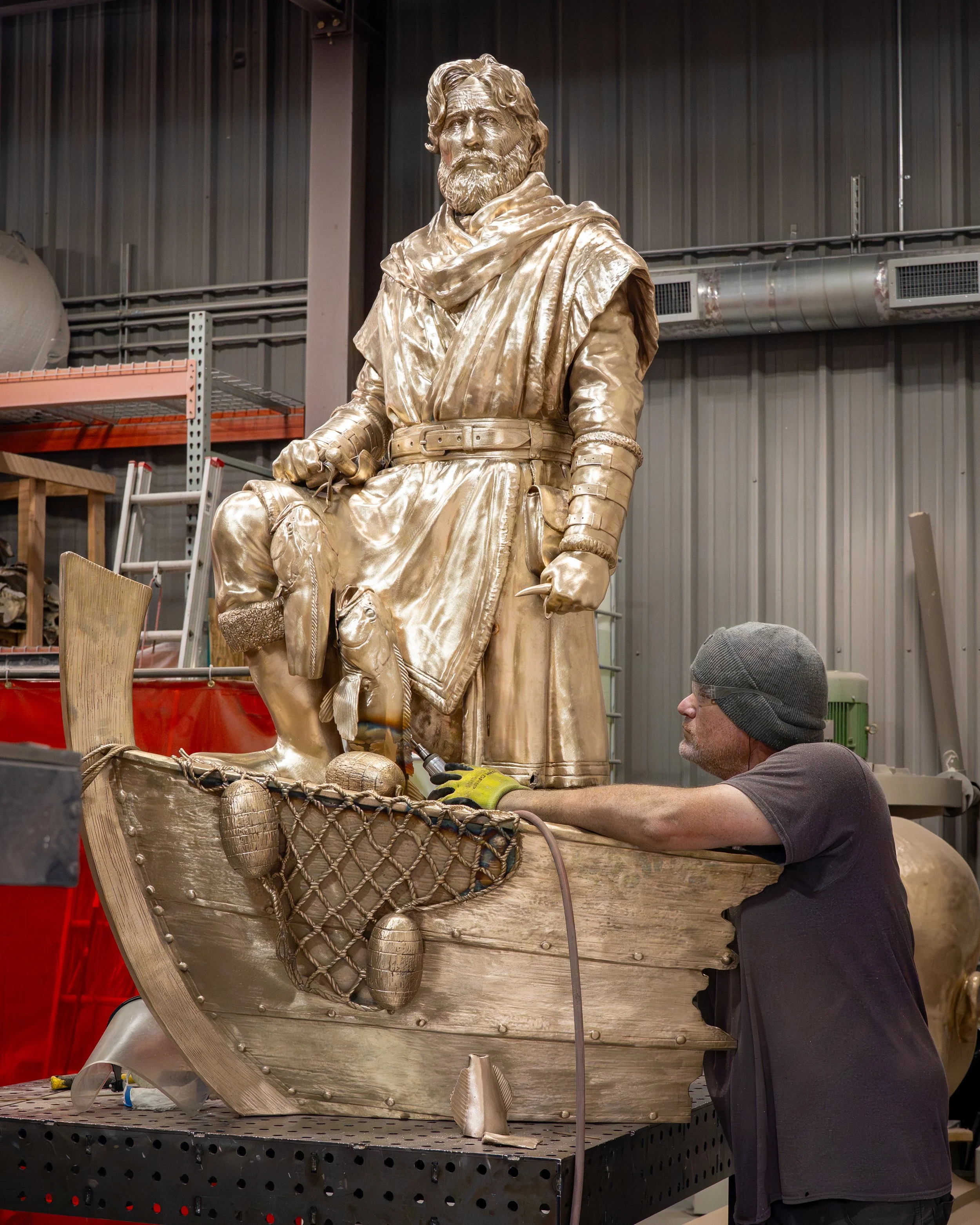 A man working on a gold-colored statue of a man with a beard, seated with one leg crossed over the other, inside a workshop with metal walls, shelves, and tools.