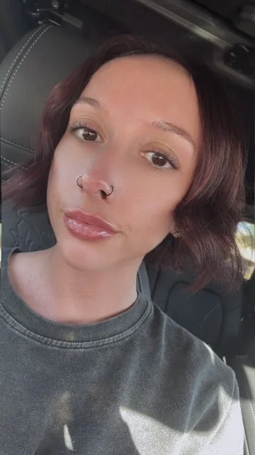 Close-up of a woman with shoulder-length dark red hair, wearing a nose ring and a grey t-shirt, sitting in a vehicle.