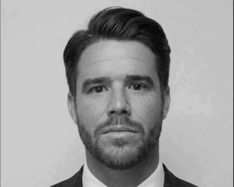 Black and white portrait of a man with neatly combed hair, beard, and mustache, wearing a collared shirt and blazer, looking directly at the camera.