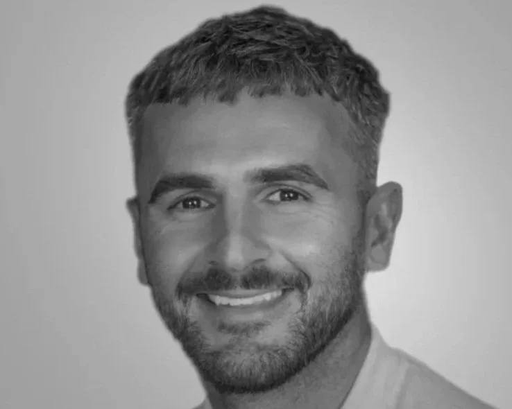 A black and white headshot of a smiling man with short hair and a beard, wearing a light-colored shirt.