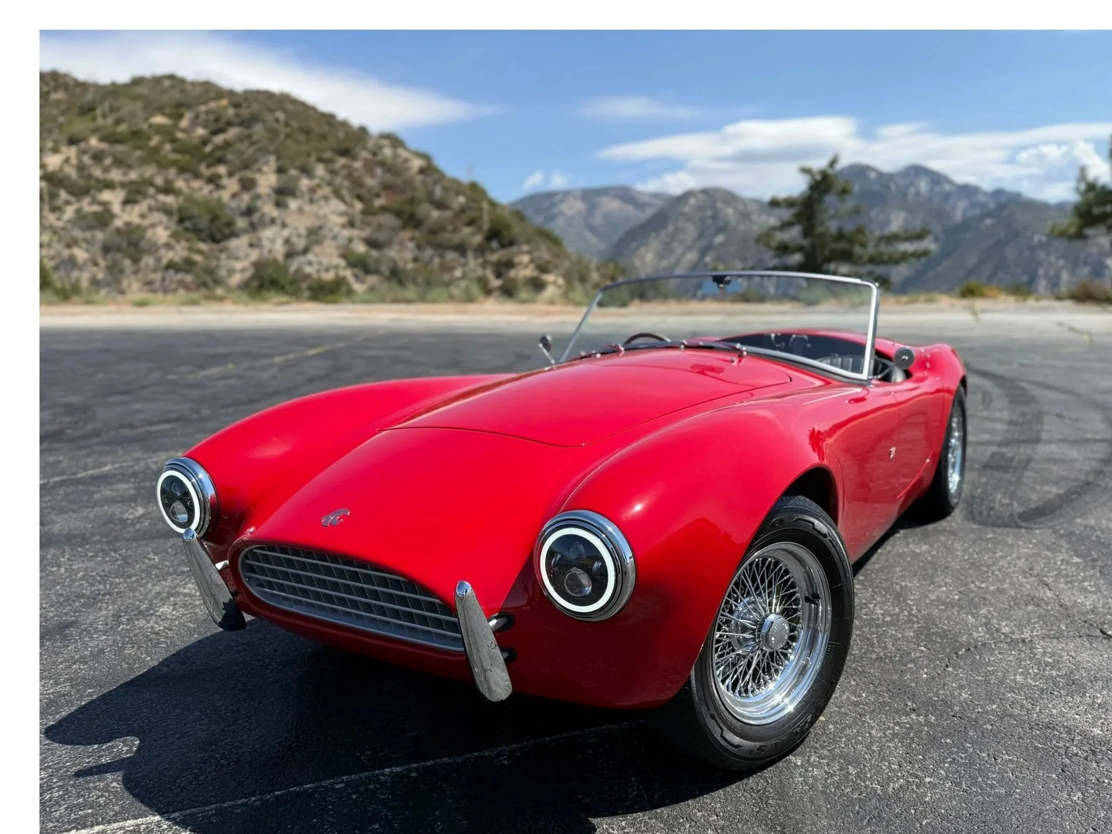 A red vintage convertible sports car parked on an asphalt road with mountains and trees in the background under a partly cloudy sky.