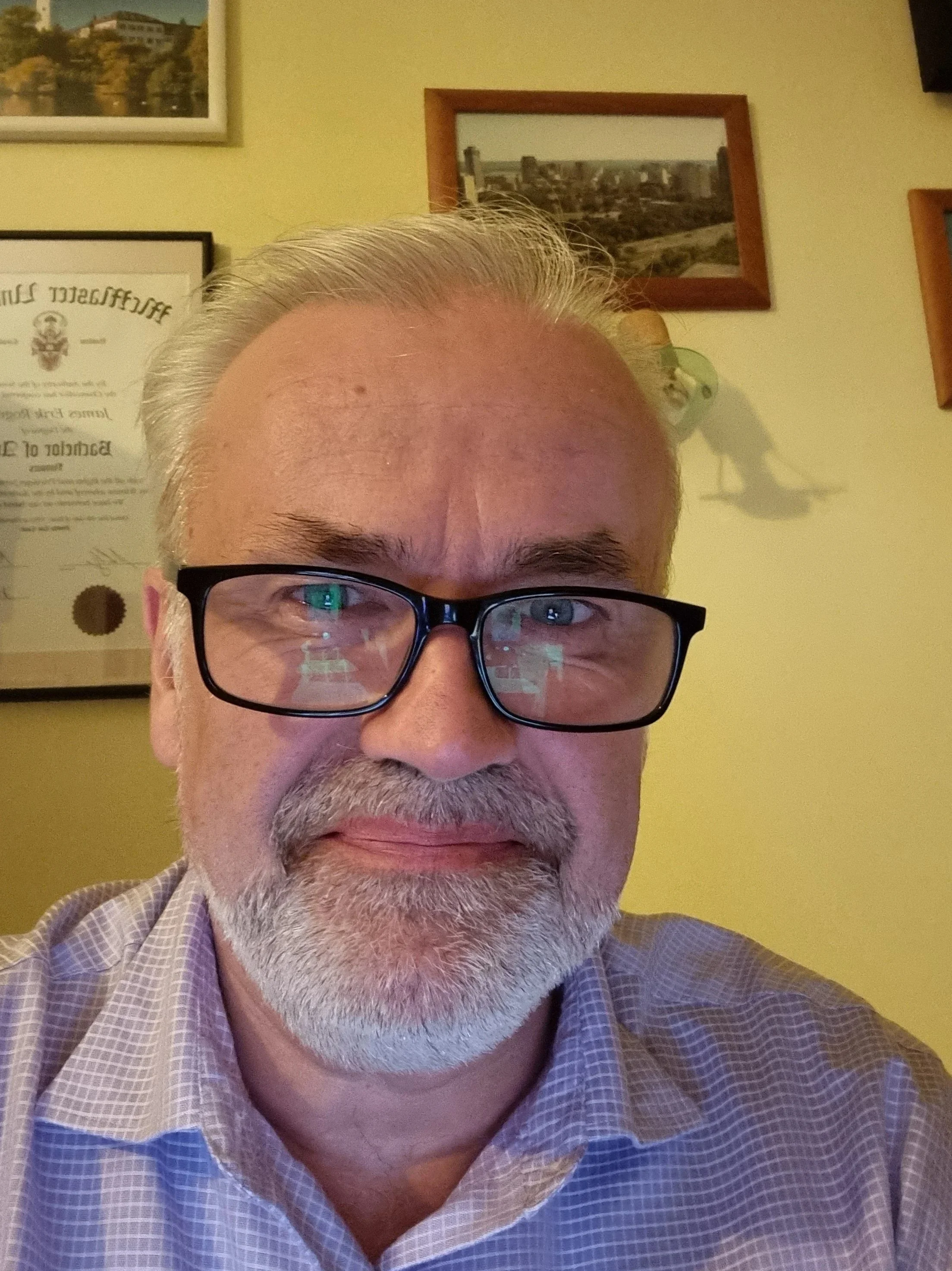 Close-up of a middle-aged man with gray hair, beard, glasses, wearing a checkered shirt, smiling slightly, with framed pictures and certificates on a yellow wall behind him.