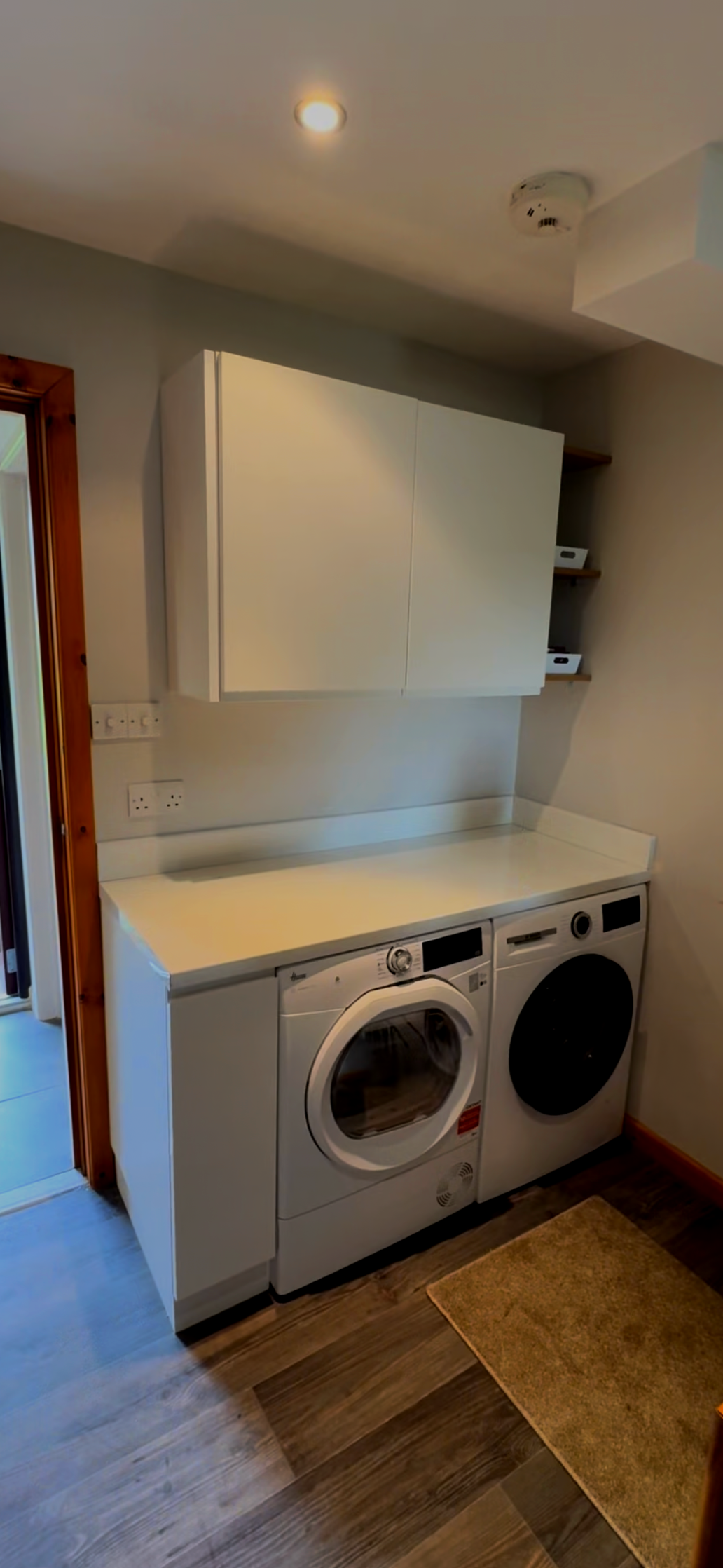 Laundry room with white front-loading washing machine and dryer, white countertop, wall-mounted white cabinet, and shelves with storage boxes. Wooden trim around door and dark wood flooring.