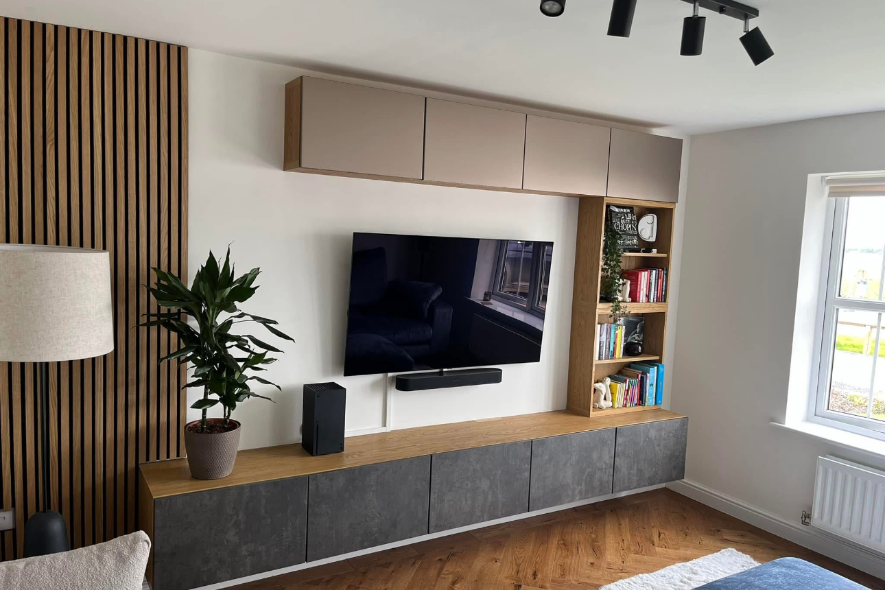 Modern living room with a flat-screen TV mounted on the wall, a bookshelf with books, a potted plant, and a window letting in natural light.