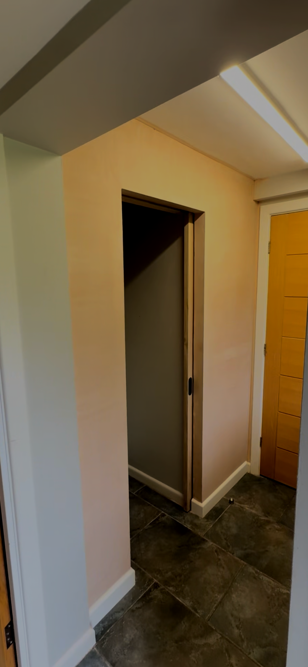 Photo of an empty elevator shaft in a hallway, with the elevator doors open and a wooden door to the right, beige and gray walls, and dark tiled floor.