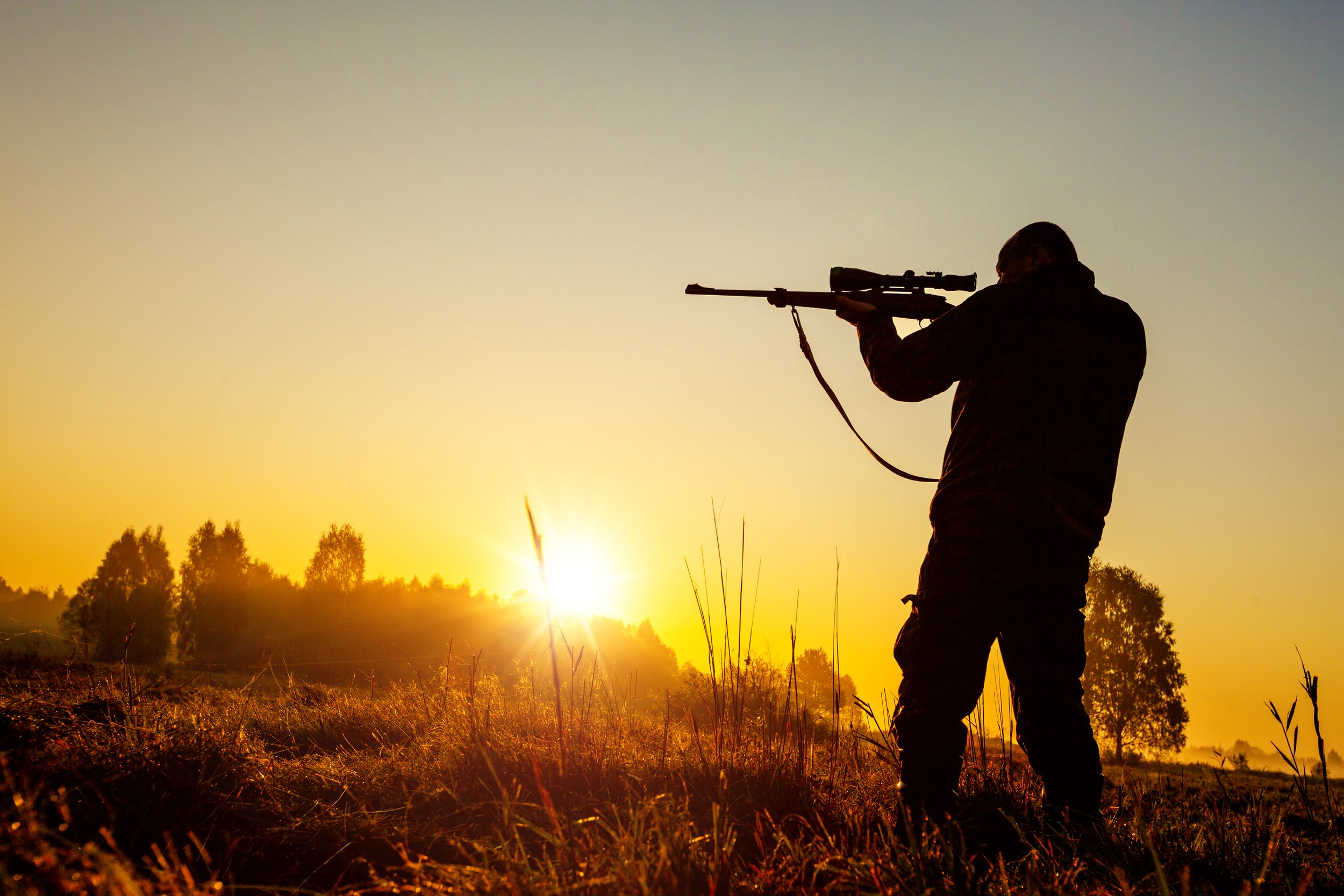 Silhouette of a man aiming a rifle backlit by a sunset.