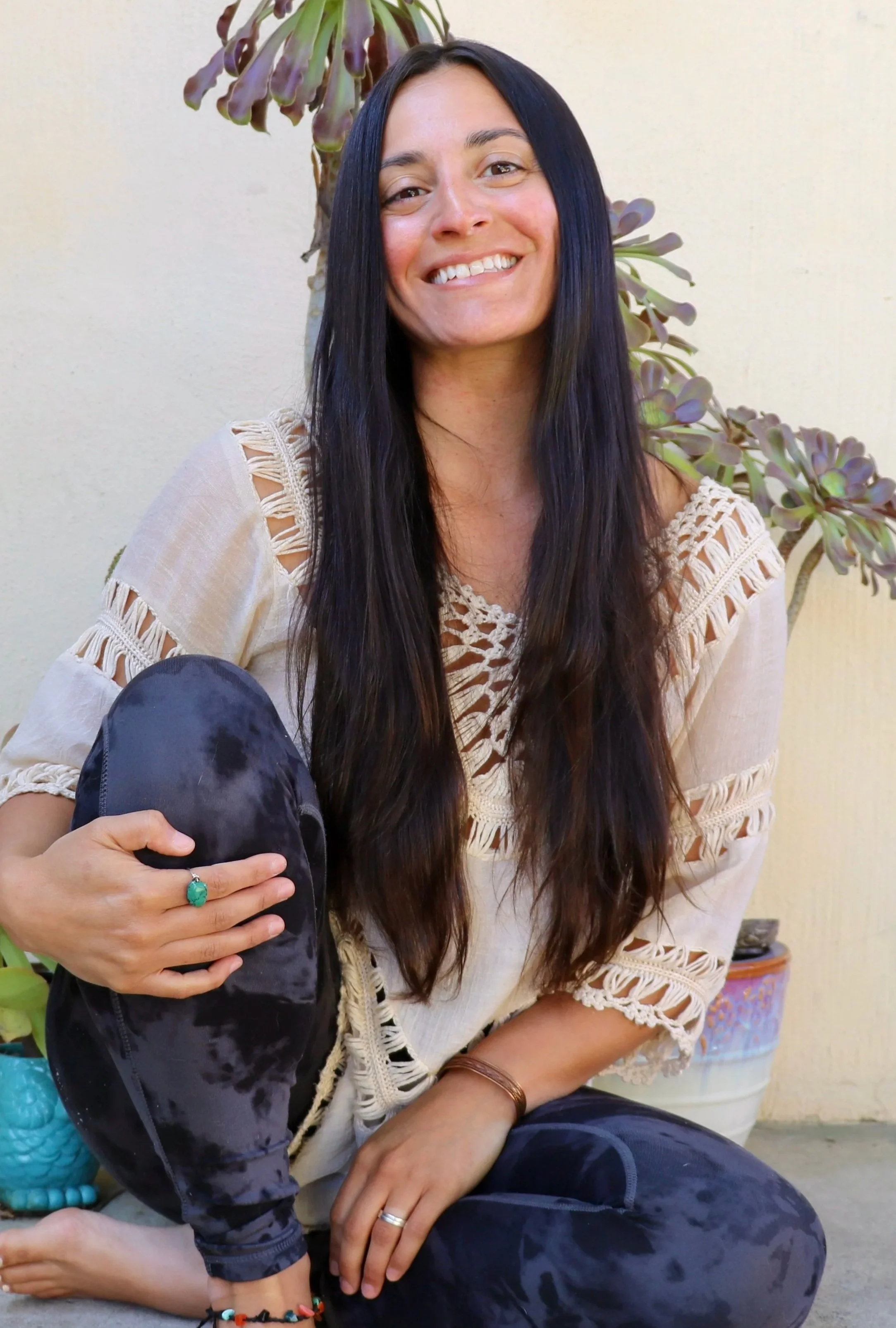 A woman with long dark hair smiling, sitting outdoors against a light-colored wall with a succulent plant in the background. She is wearing a beige crochet top, patterned pants, and a turquoise ring on her finger, holding a black tie-dye hoodie.