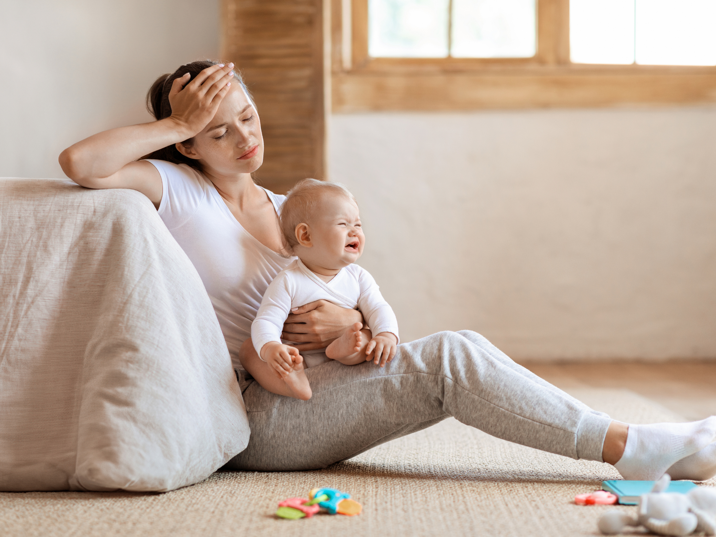 A woman sitting on a couch, holding a crying baby on her lap, with a window in the background. The woman appears distressed and the baby is upset.