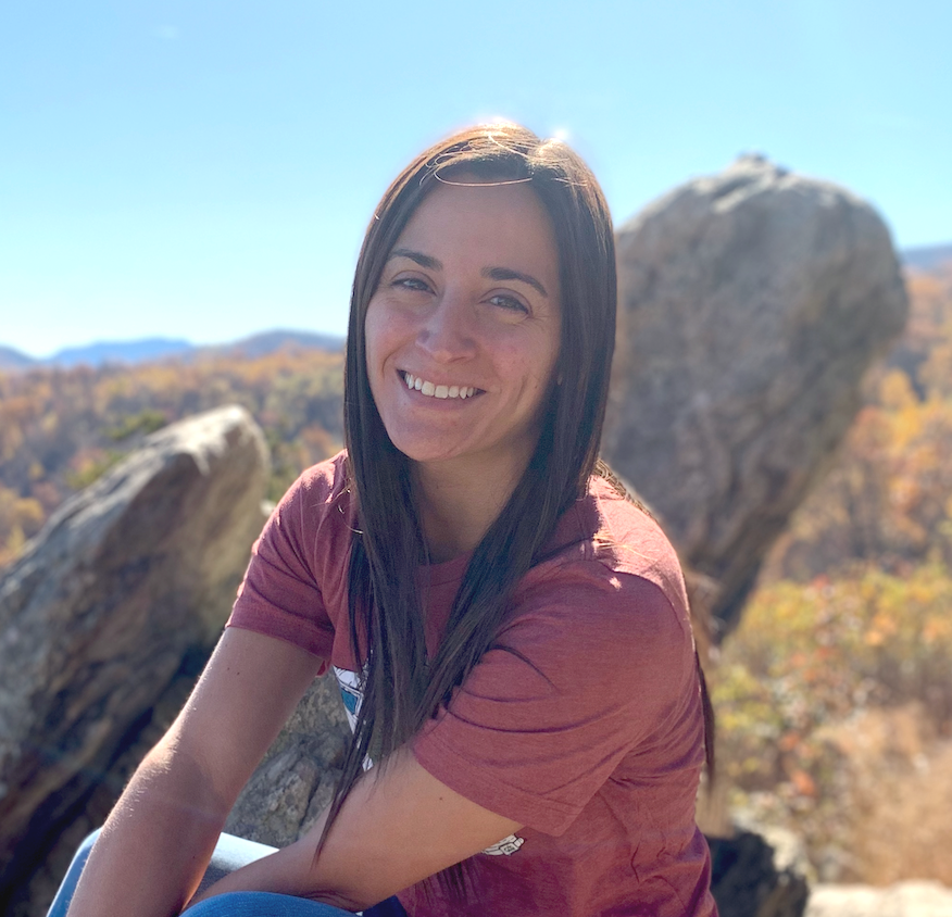 A woman with long dark hair smiling outdoors, with large rocks and trees in the background on a sunny day.