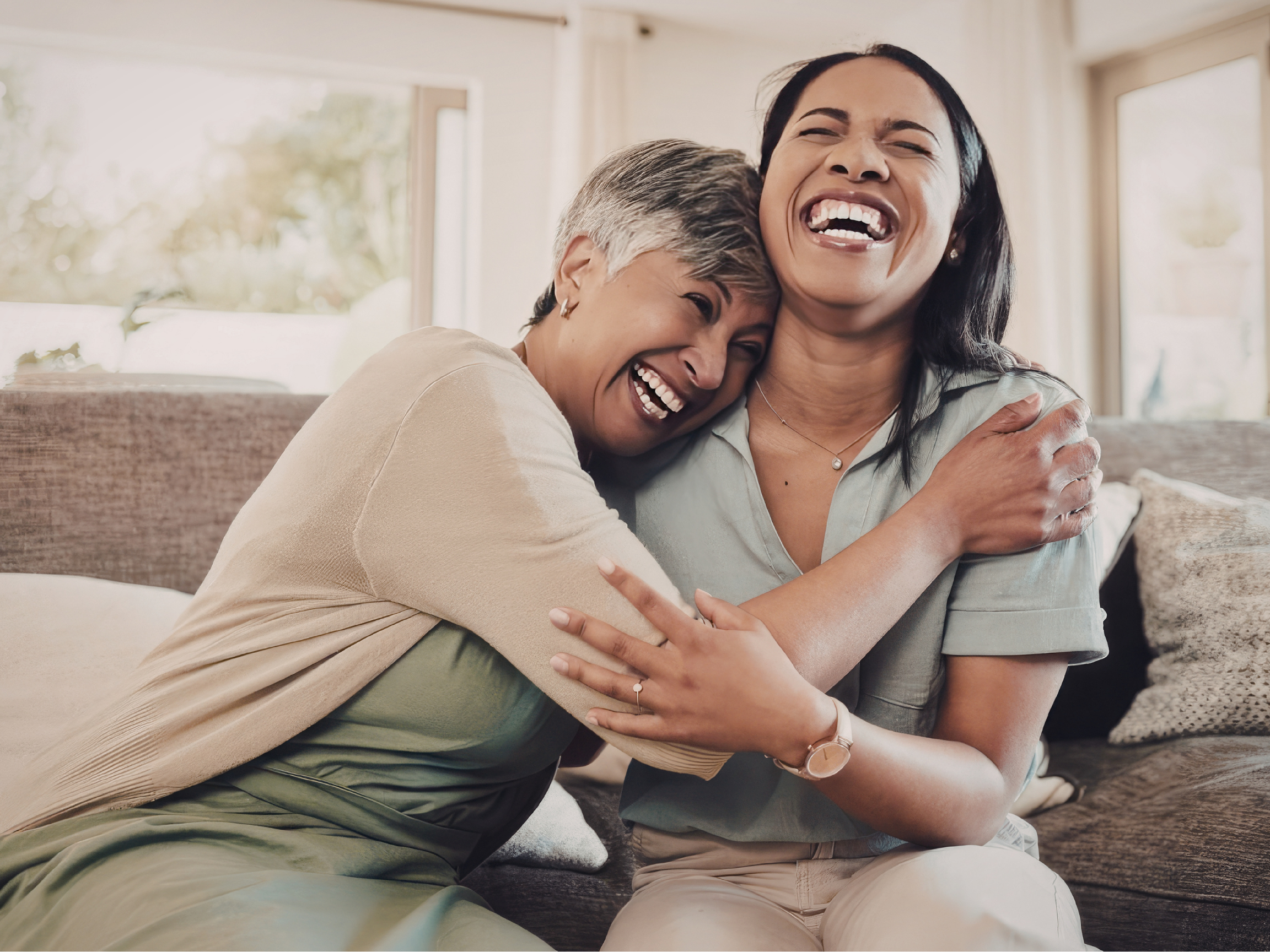 Two women, one with short gray hair and the other with long dark hair, are hugging and laughing together in a bright, cozy living room with large windows in the background.