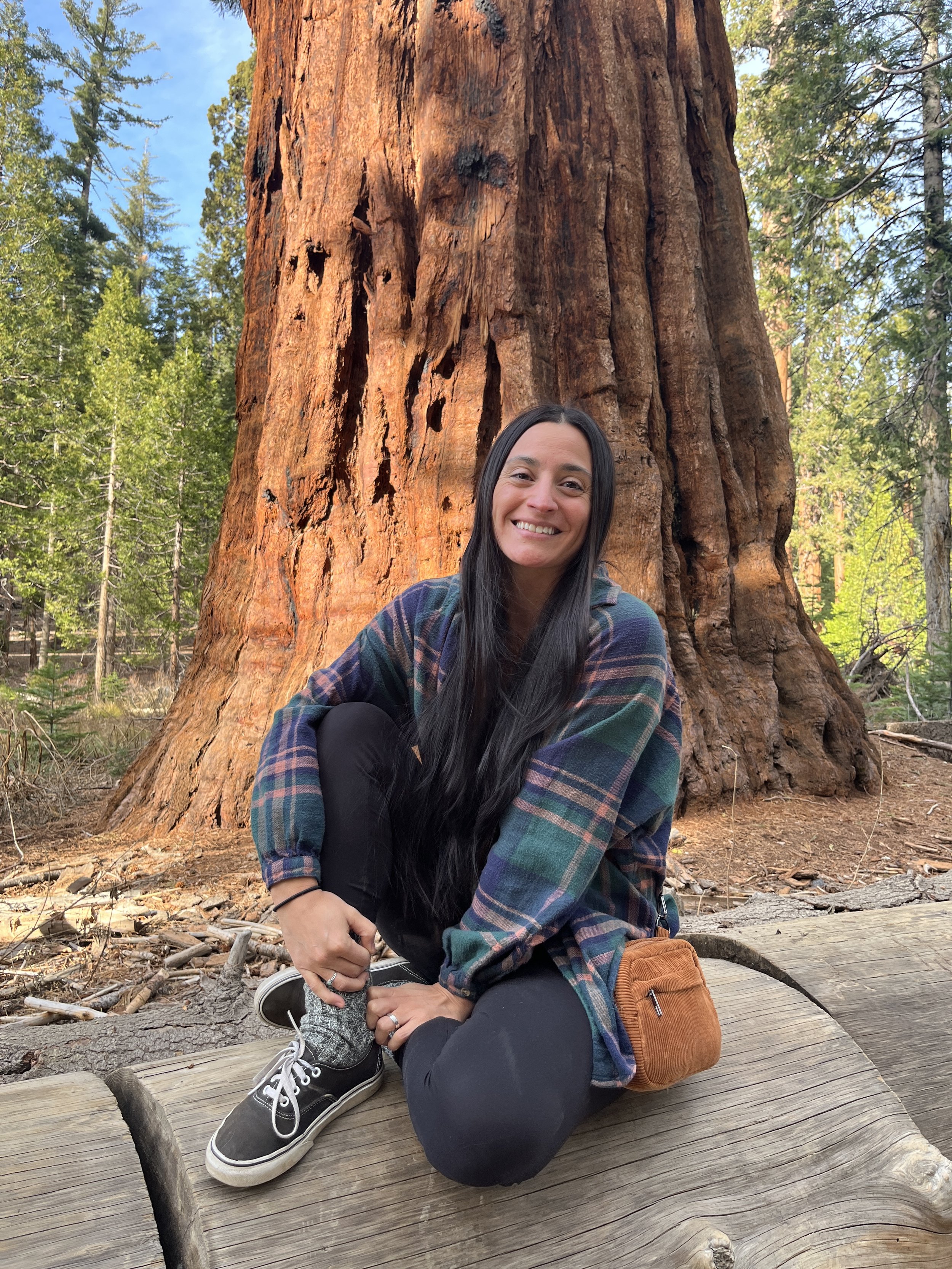 A woman with long dark hair smiling while sitting on a wooden platform in a forest, with a large redwood tree behind her.