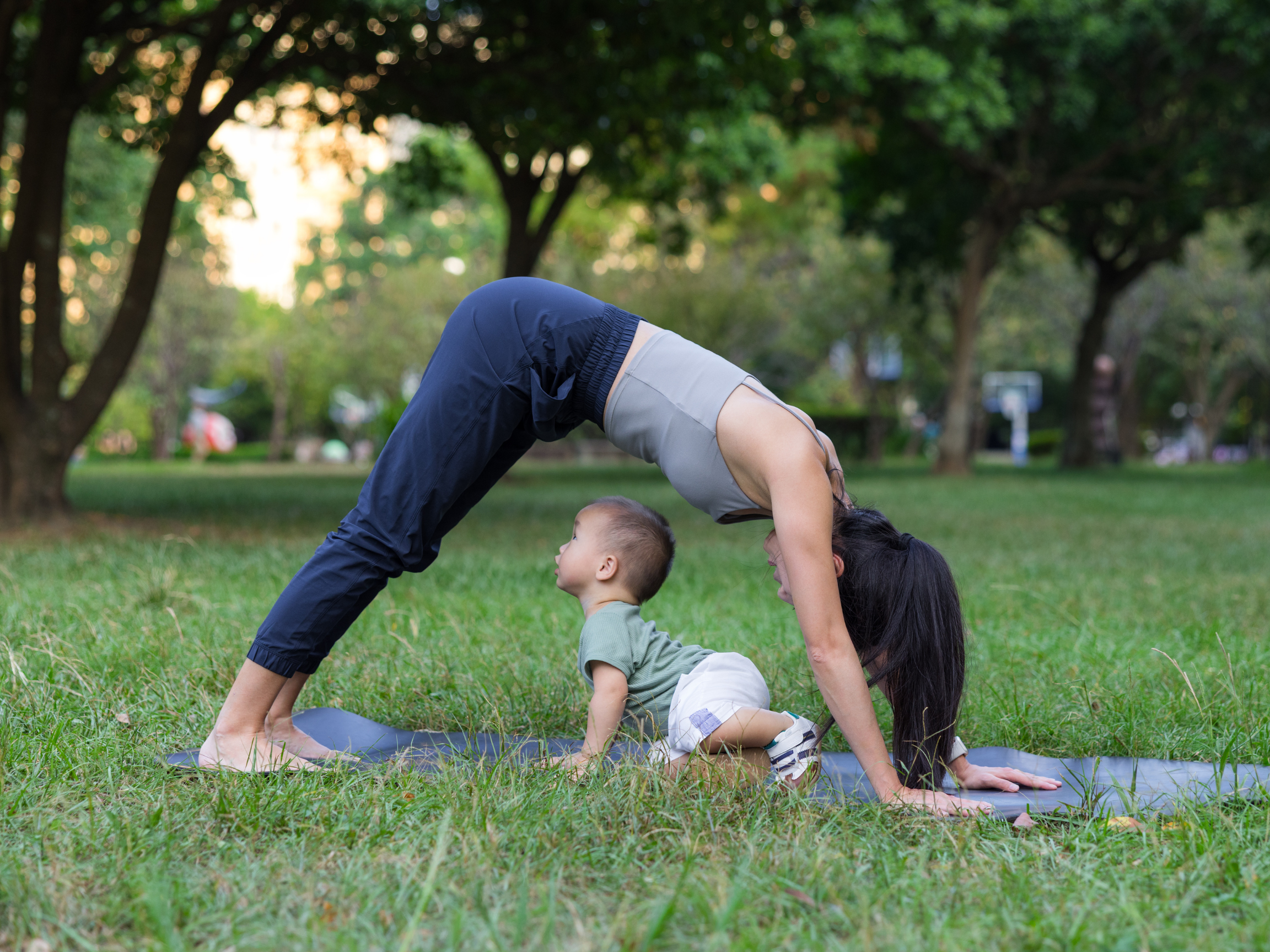 A woman and two young children practicing yoga outdoors in a park during sunset, with trees and green grass surrounding them.