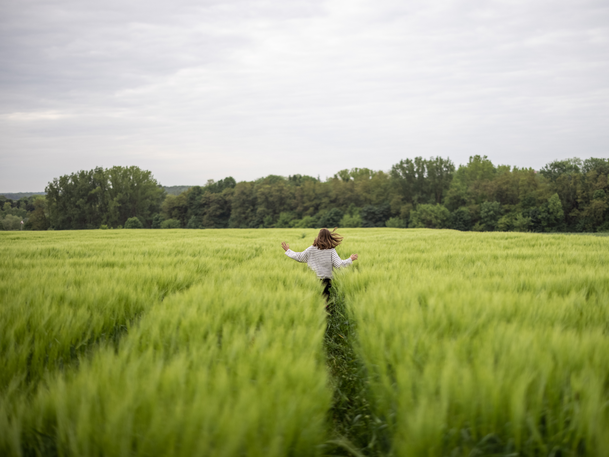 A young girl running through a green field of tall grass with a backdrop of trees and cloudy sky.