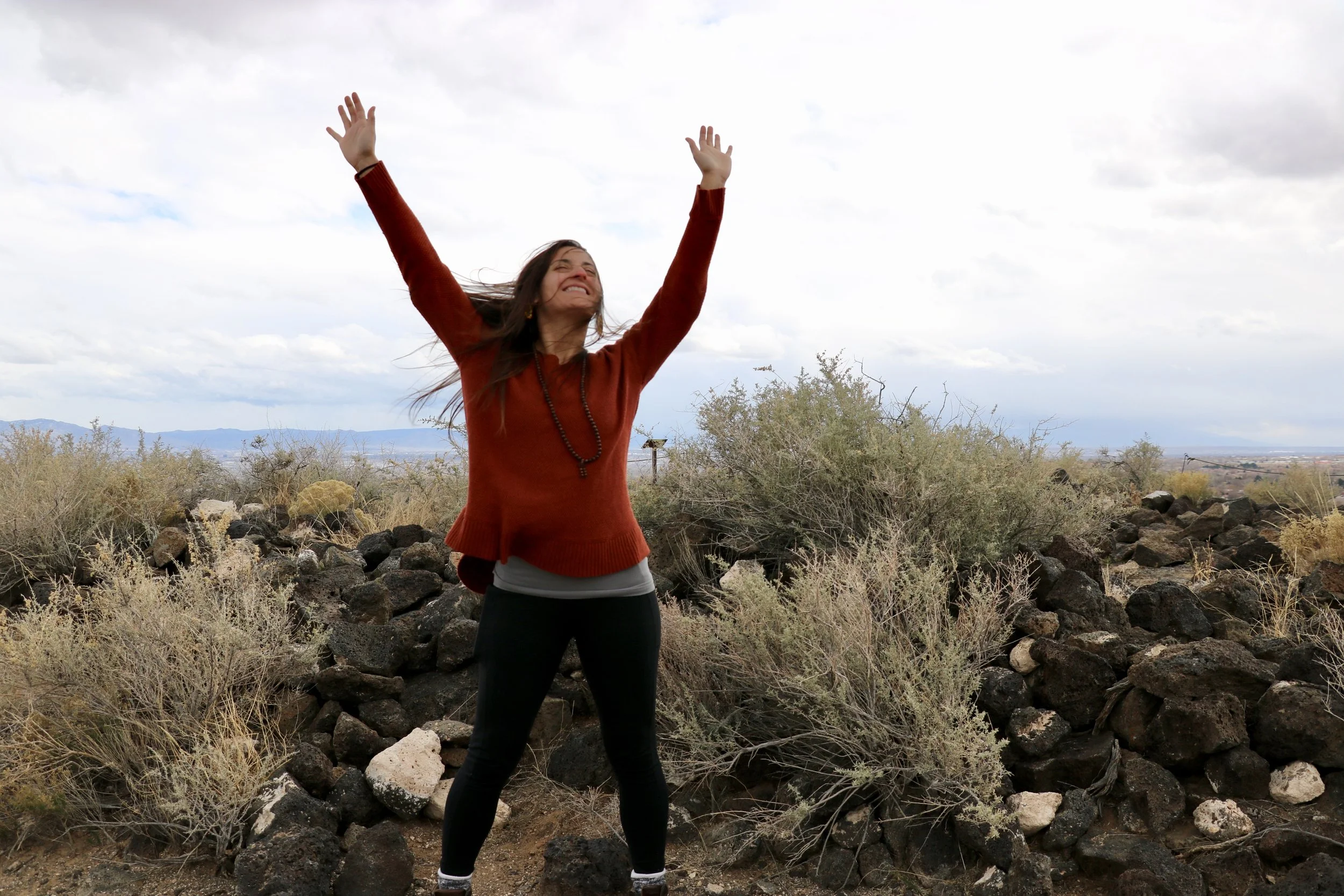 A woman standing outdoors with arms raised and smiling joyfully, surrounded by rocks and bushes under a cloudy sky.