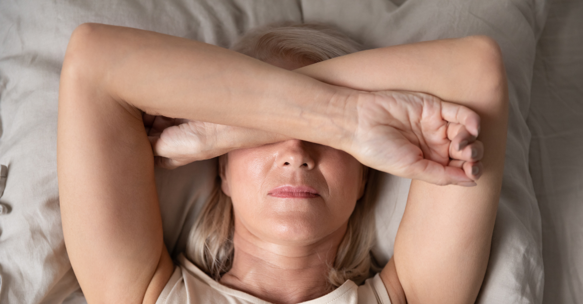 An older woman lying in bed with her arm covering her face.