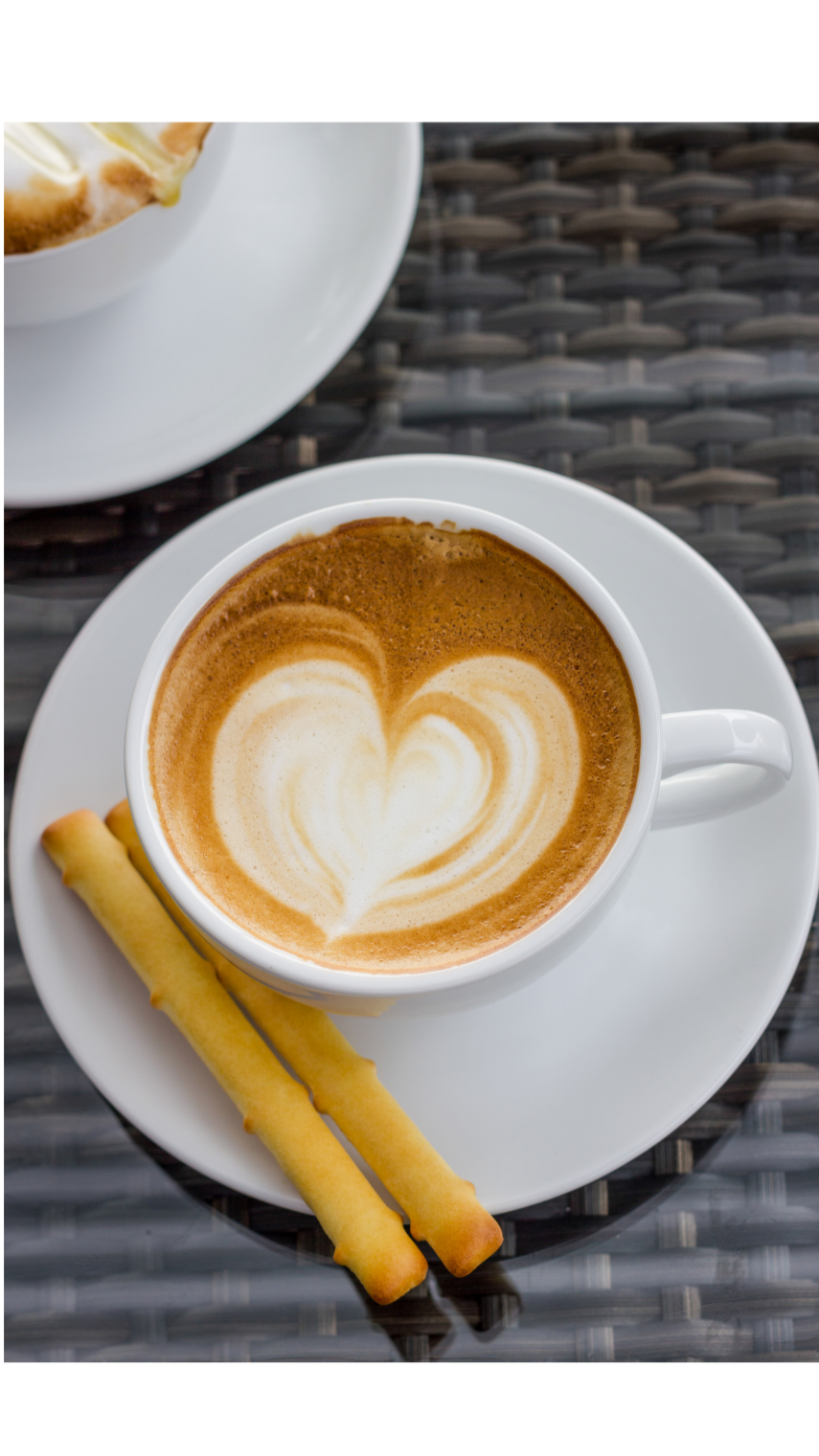 A cup of coffee with latte art in a white cup on a white saucer, with two rolled wafer cookies on the saucer. Part of another similar coffee cup with latte art is visible in the background.