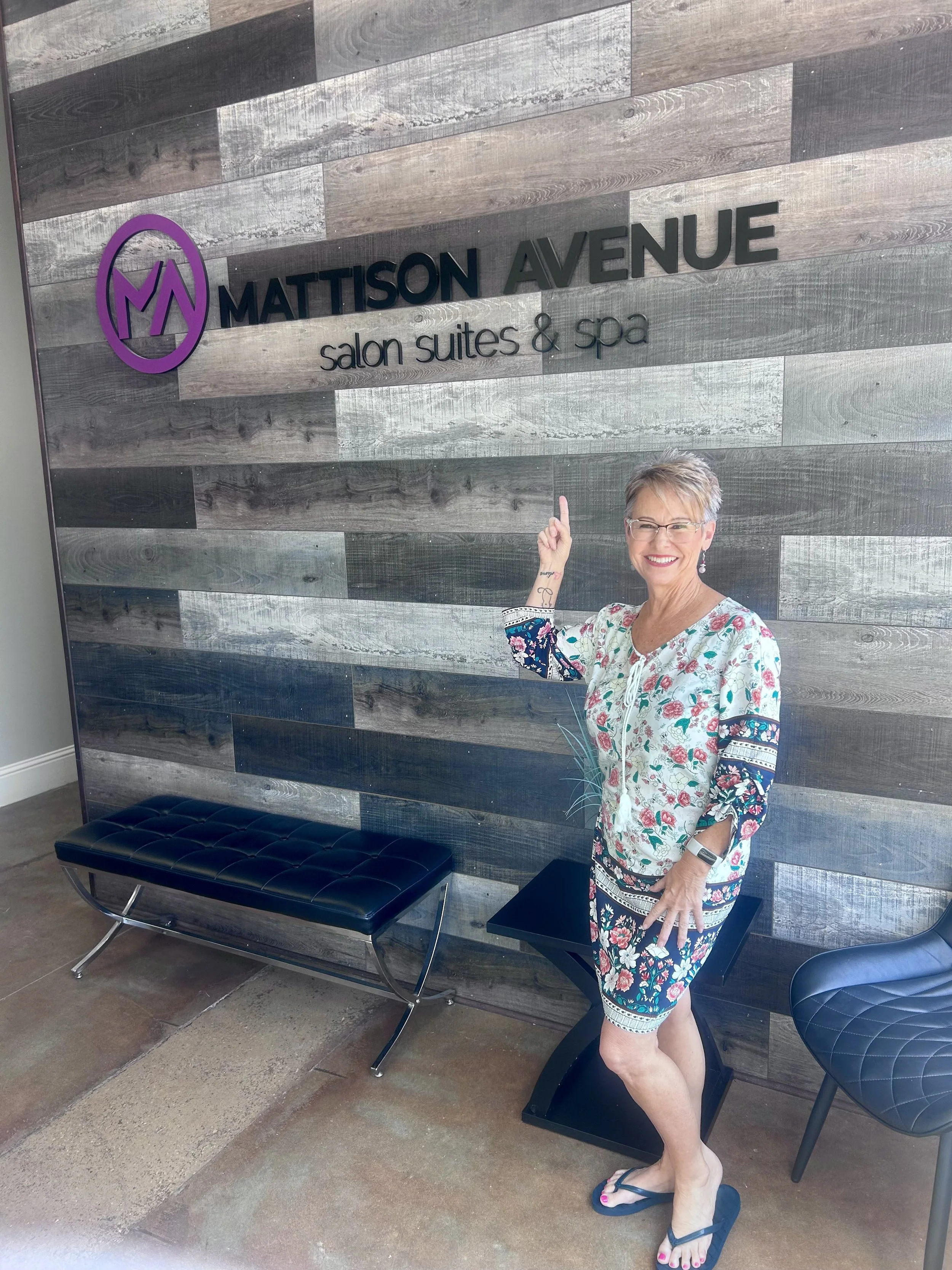 A woman with short gray hair, glasses, and a cheerful smile is standing indoors in front of a wooden wall with a sign that reads 'Mattison Avenue salon suites & spa.' She is wearing a colorful floral dress, flip-flops, and has a tattoo on her wrist. She is pointing up with her right index finger.