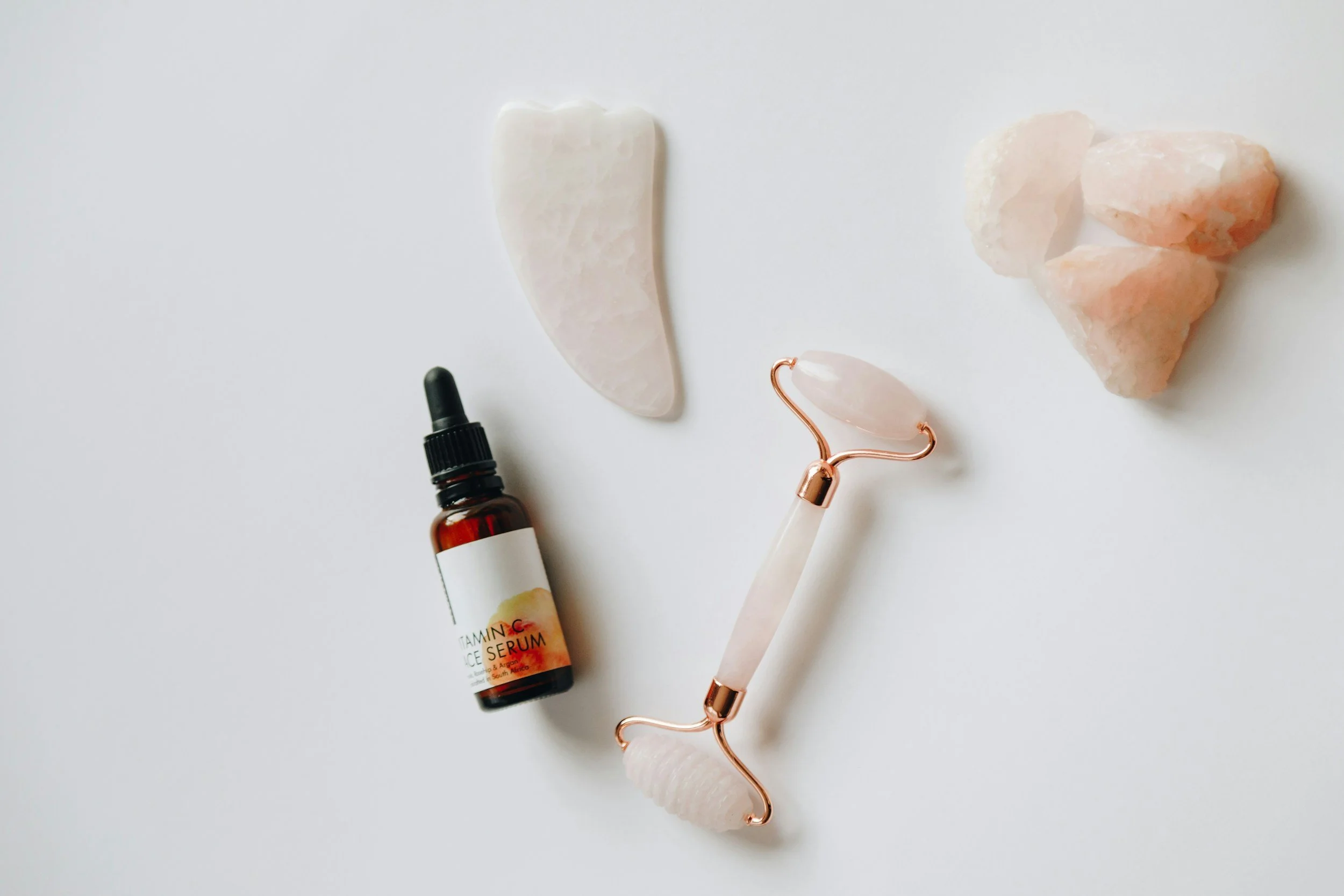 Skincare tools and products on a white surface, including a jade roller, rose quartz face roller, salt stones, essential serum bottle, and a black dropper.