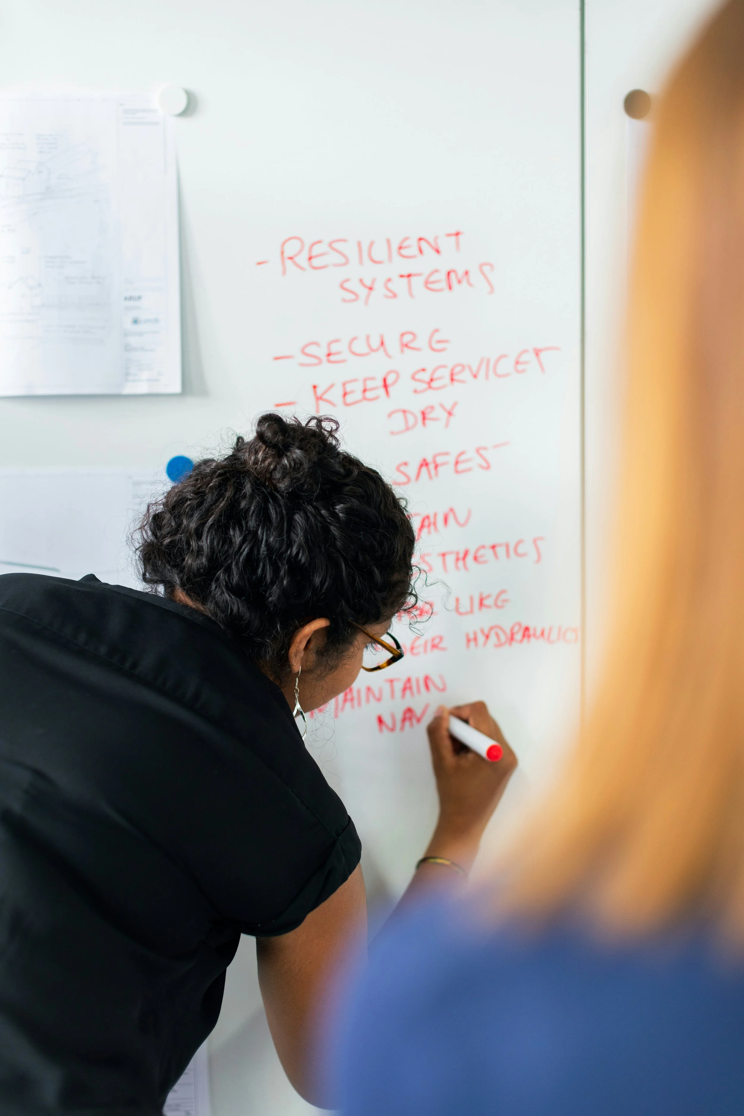 A woman, wearing glasses and a black shirt, writing on a whiteboard with a red marker. The whiteboard has handwritten notes in red ink, including words like 'Resilient Systems,' 'Secure,' and 'Keep Service Dry.'