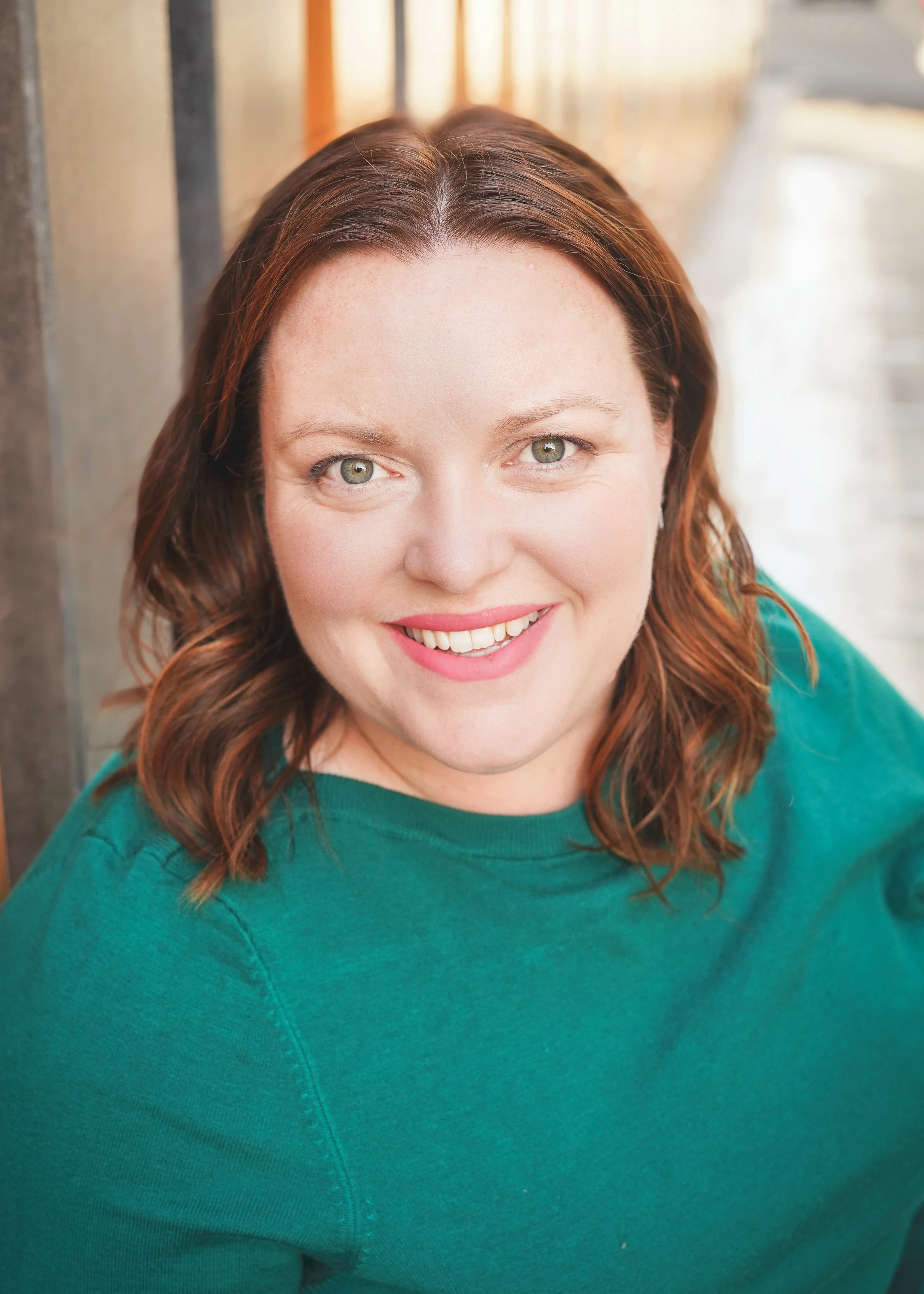 Close-up of woman with reddish-brown hair, green eyes, smiling in a teal sweater, outdoors with blurred background.