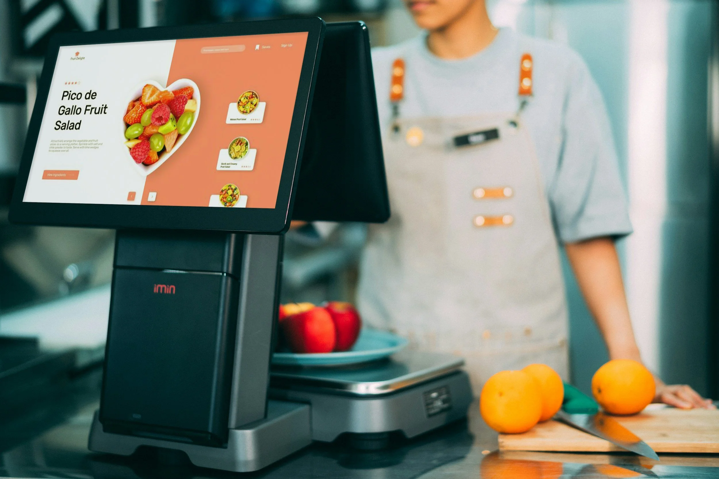 Close-up of a digital cash register screen displaying a fruit salad recipe titled 'Pico de Gallo Fruit Salad' with an image of a fruit salad and recipe options on the display, in a grocery store or market setting.