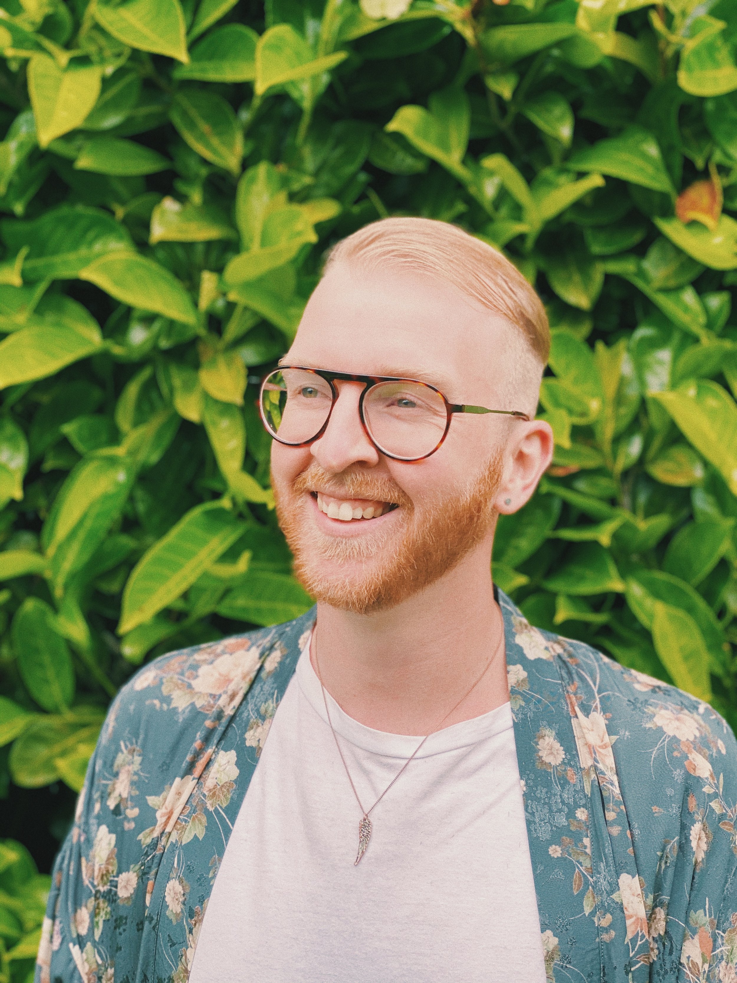 Photo of a smiling man with red hair and a beard, wearing glasses, a white t-shirt, a floral shirt, and a necklace, standing outdoors in front of green foliage.