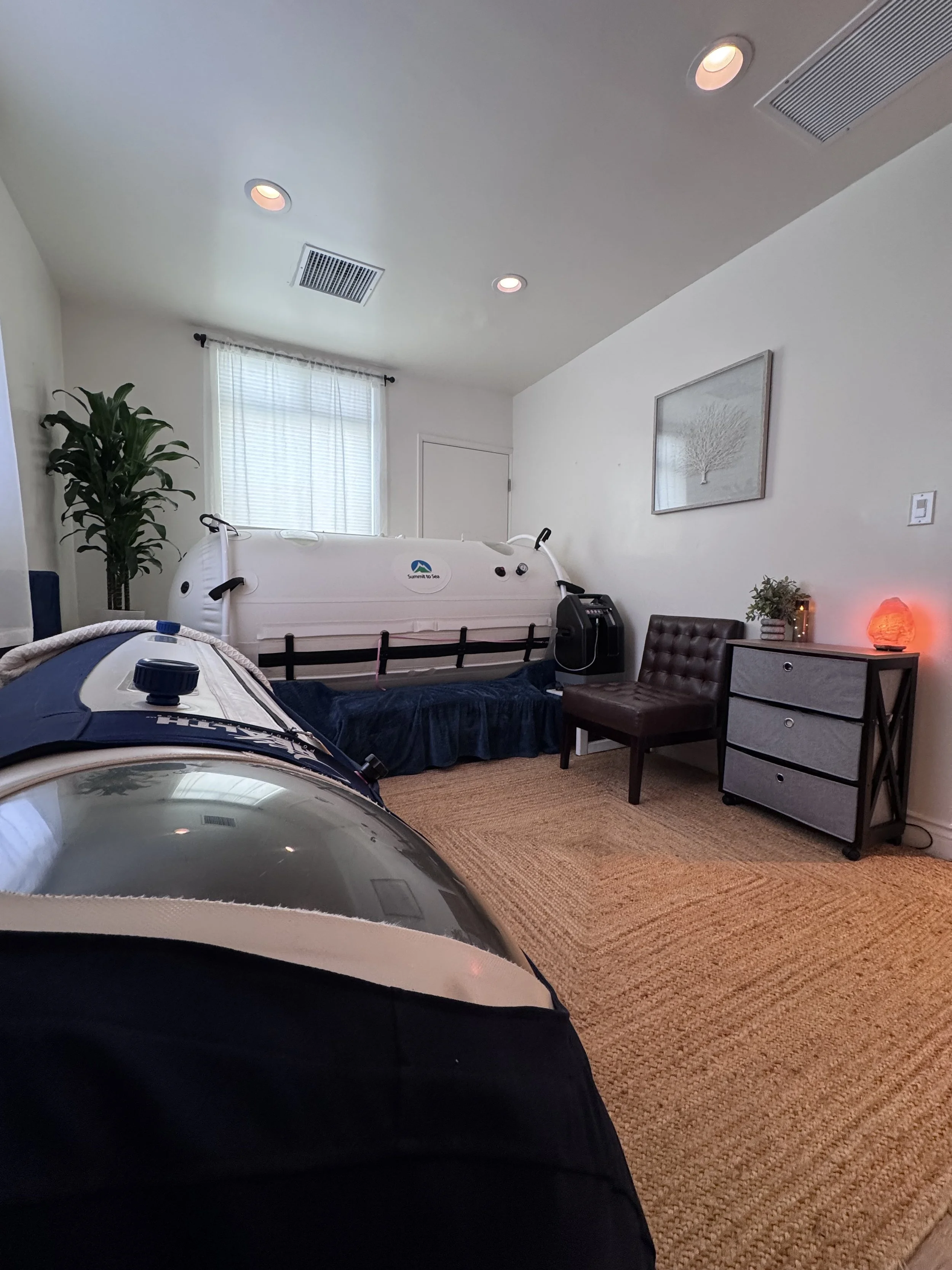 Room with 2 hyperbaric oxygen chambers, a potted plant, and medical equipment, indicating a sensory or relaxation space.