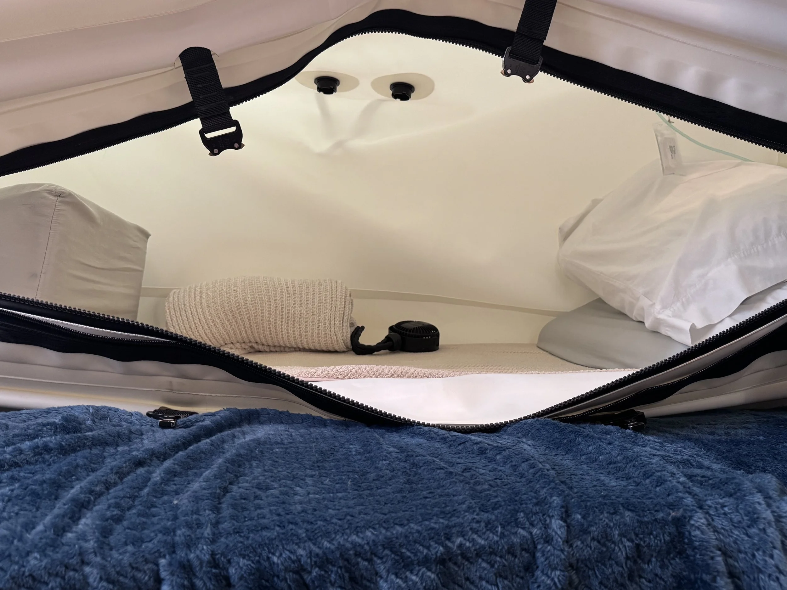 View of inside a hyperbaric chamber, showing pillows , a beige blanket, and some white bedding. . The photo is taken from a low angle, with a blue textured surface at the bottom of the image.