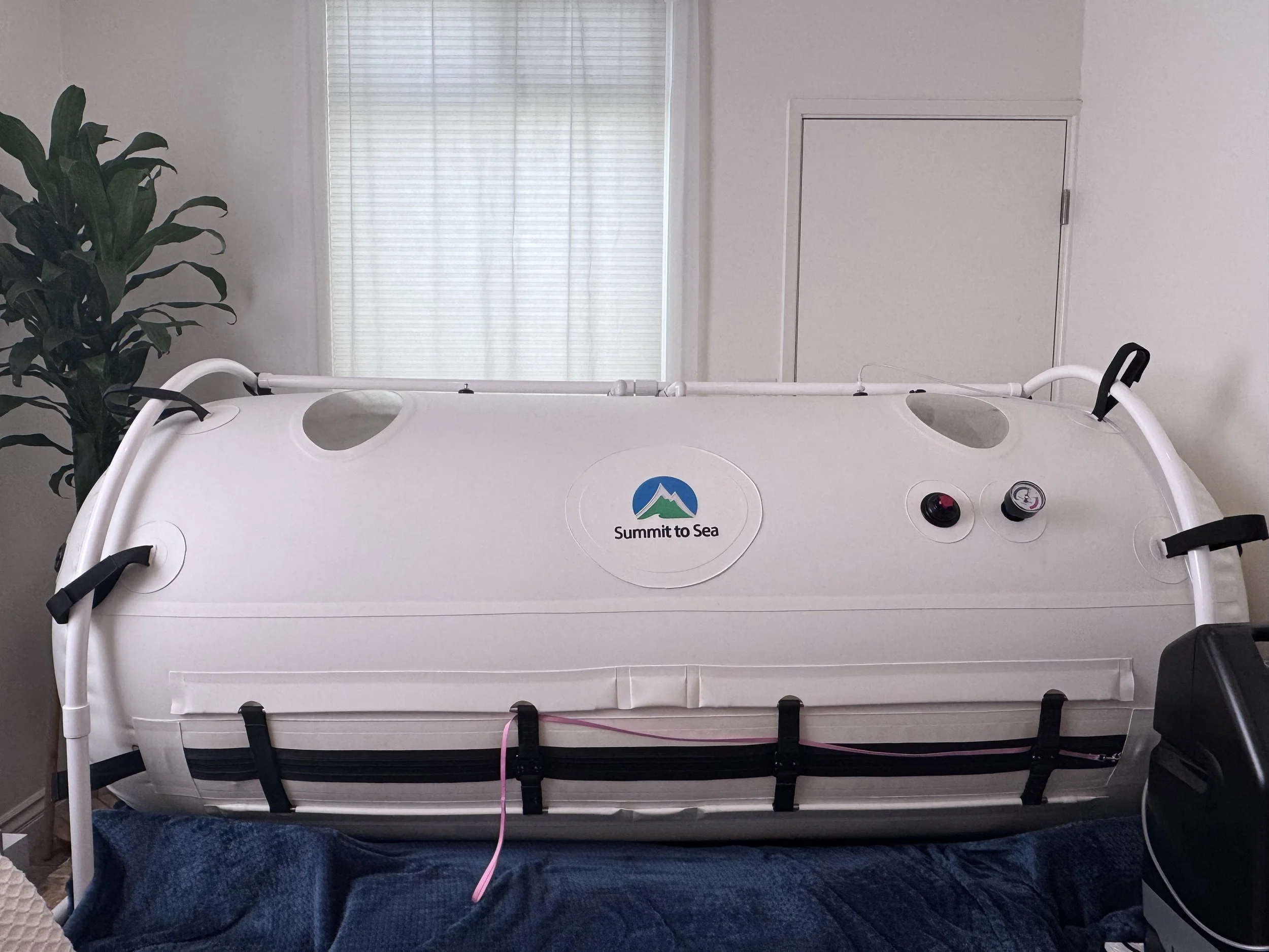 A hyperbaric oxygen chamber placed in a room with light-colored walls, a window with curtains, and a potted plant to the side.
