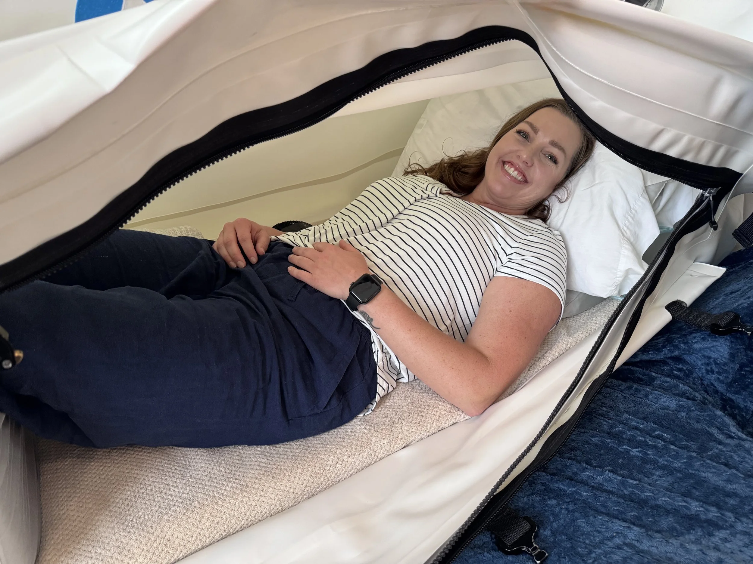 A woman lying a hyperbaric oxygen chamber, smiling and looking at the camera, wearing a striped shirt and a smartwatch.