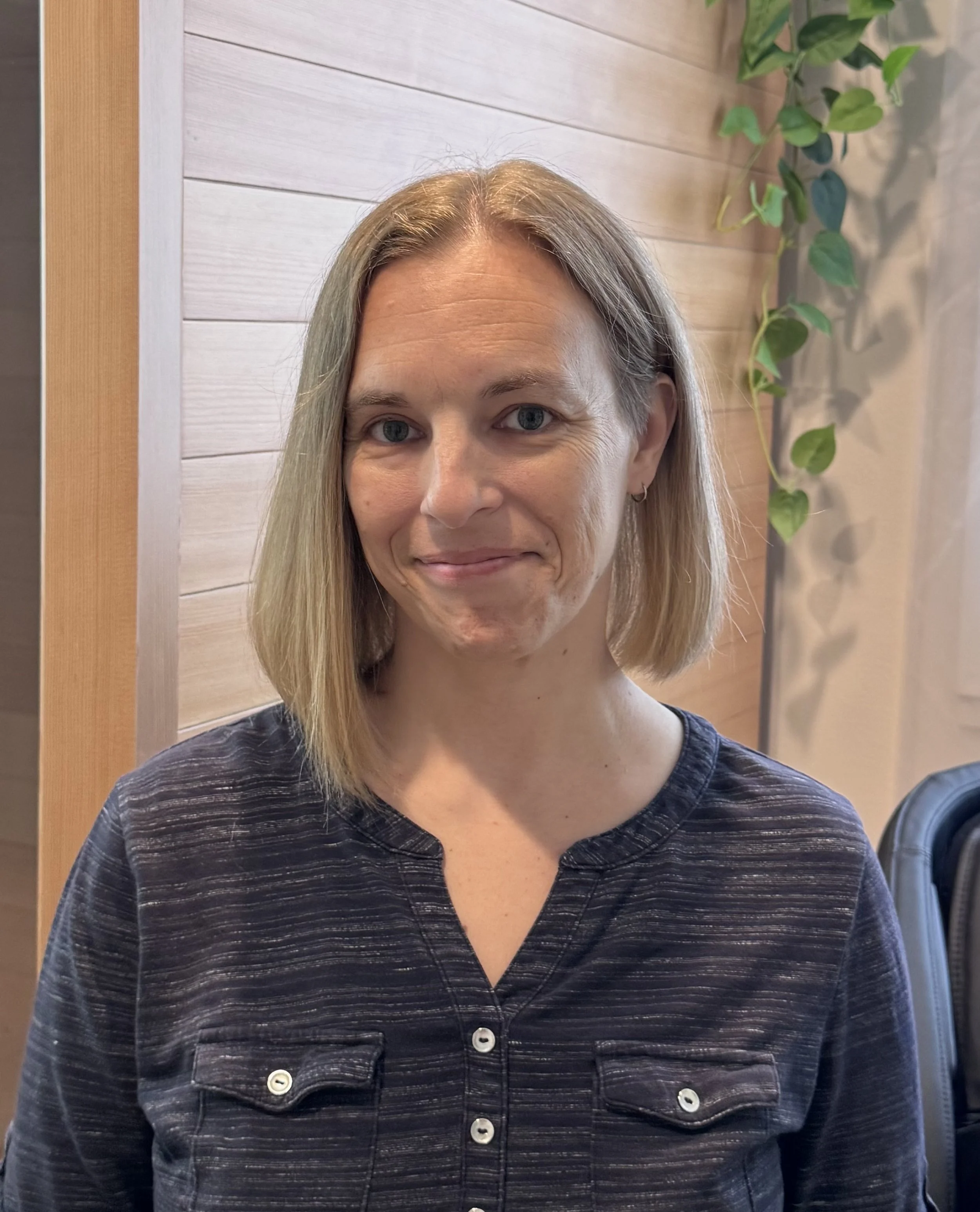 A woman with blonde hair and blue eyes smiling, wearing a dark striped shirt with buttons, indoors with wooden wall paneling and green foliage in the background.
