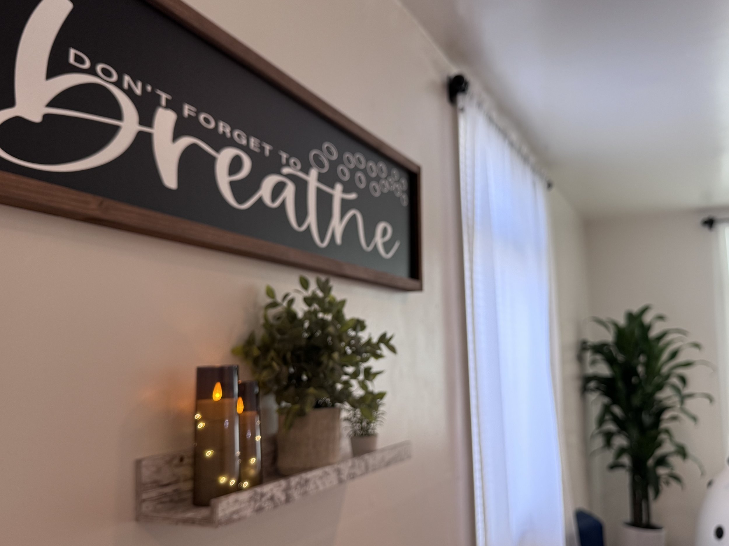 Decorative sign on wall that reads 'Don't forget to breathe'. Items on a mantle, including a potted plant and candles, are visible below the sign. Bright window with sheer curtains in the background.