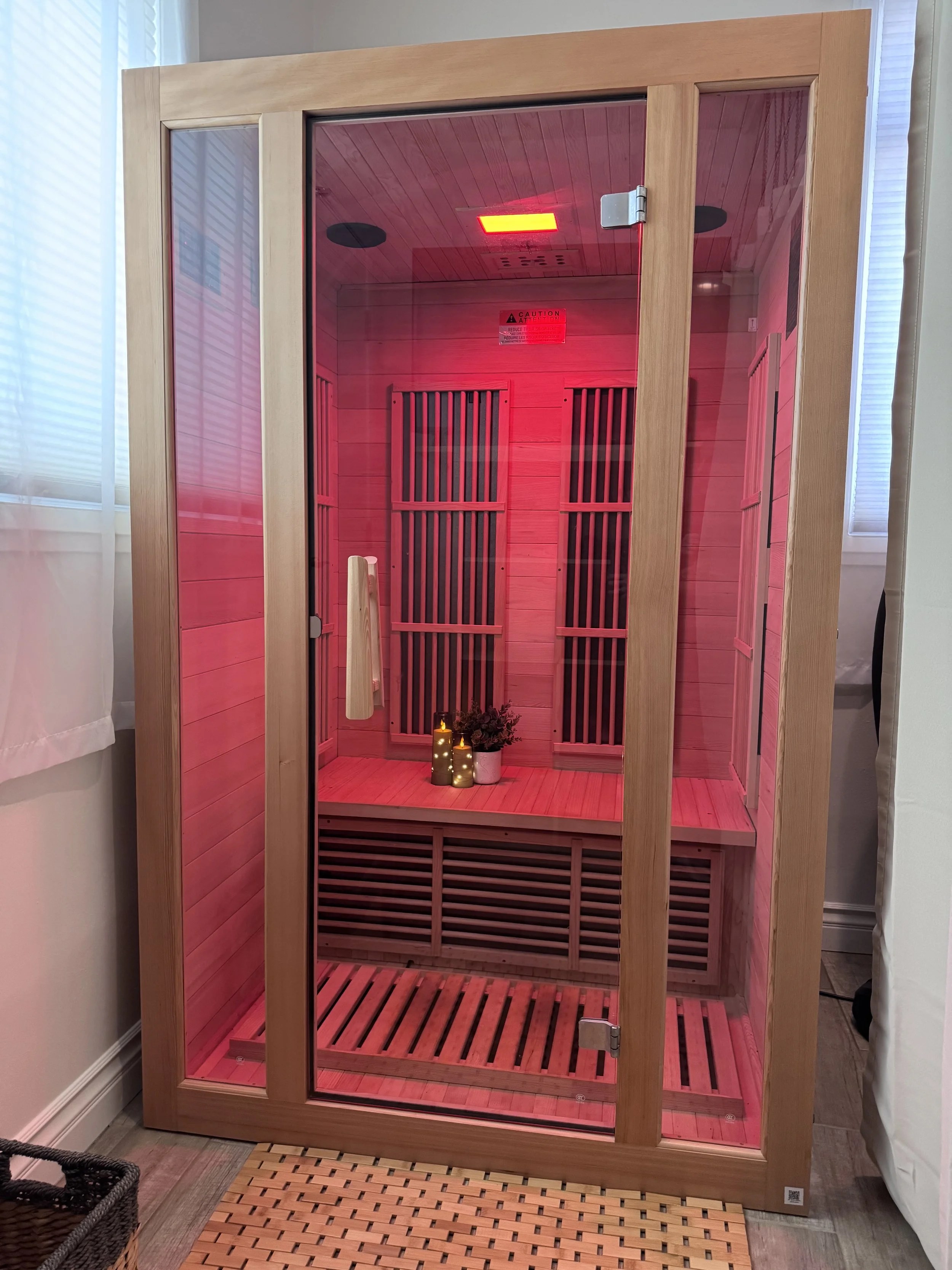 An indoor infrared sauna with a glass door and wooden interior, red lighting inside, candles and plants on the floor.