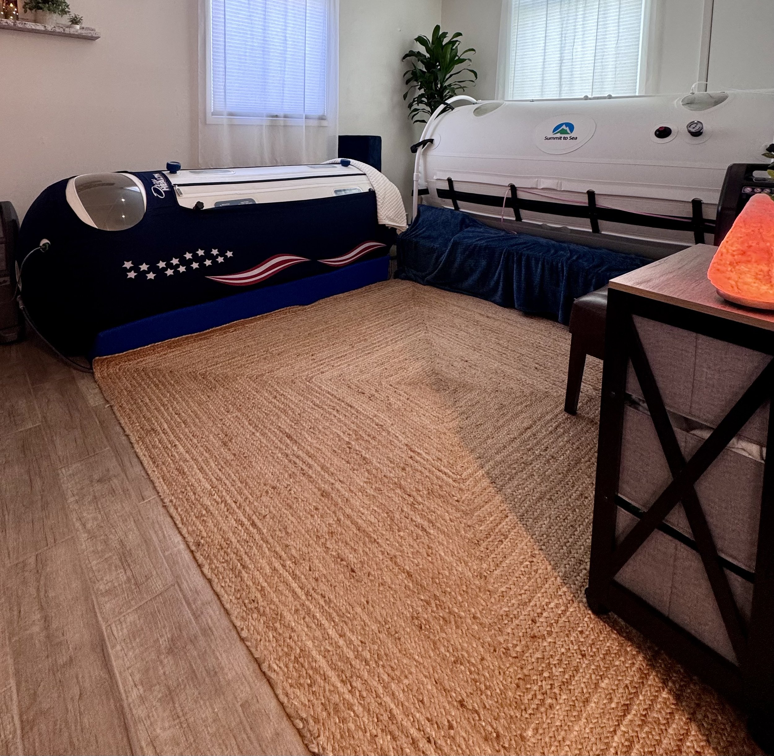 Two hyperbaric oxygen chambers in a room with beige walls, wood flooring, a plant, and windows with blinds.