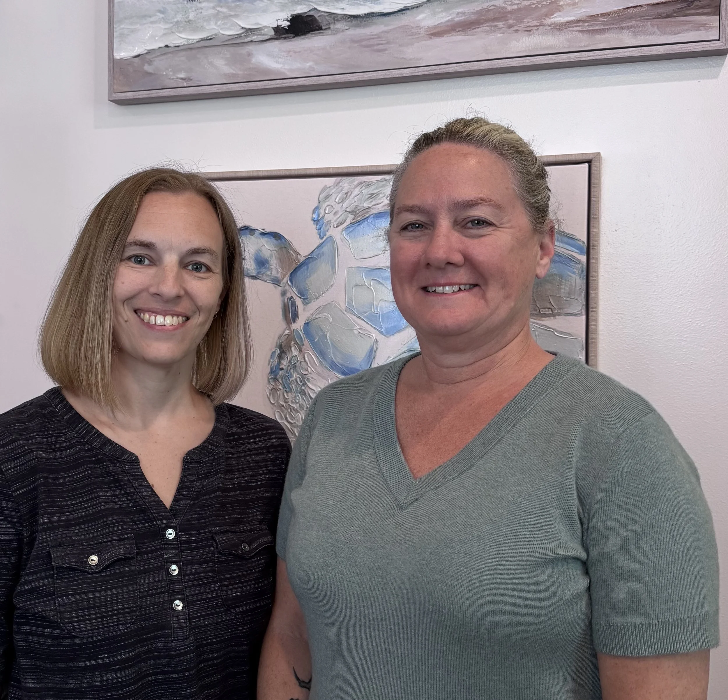 Two women smiling, standing in front of abstract artwork on white wall.