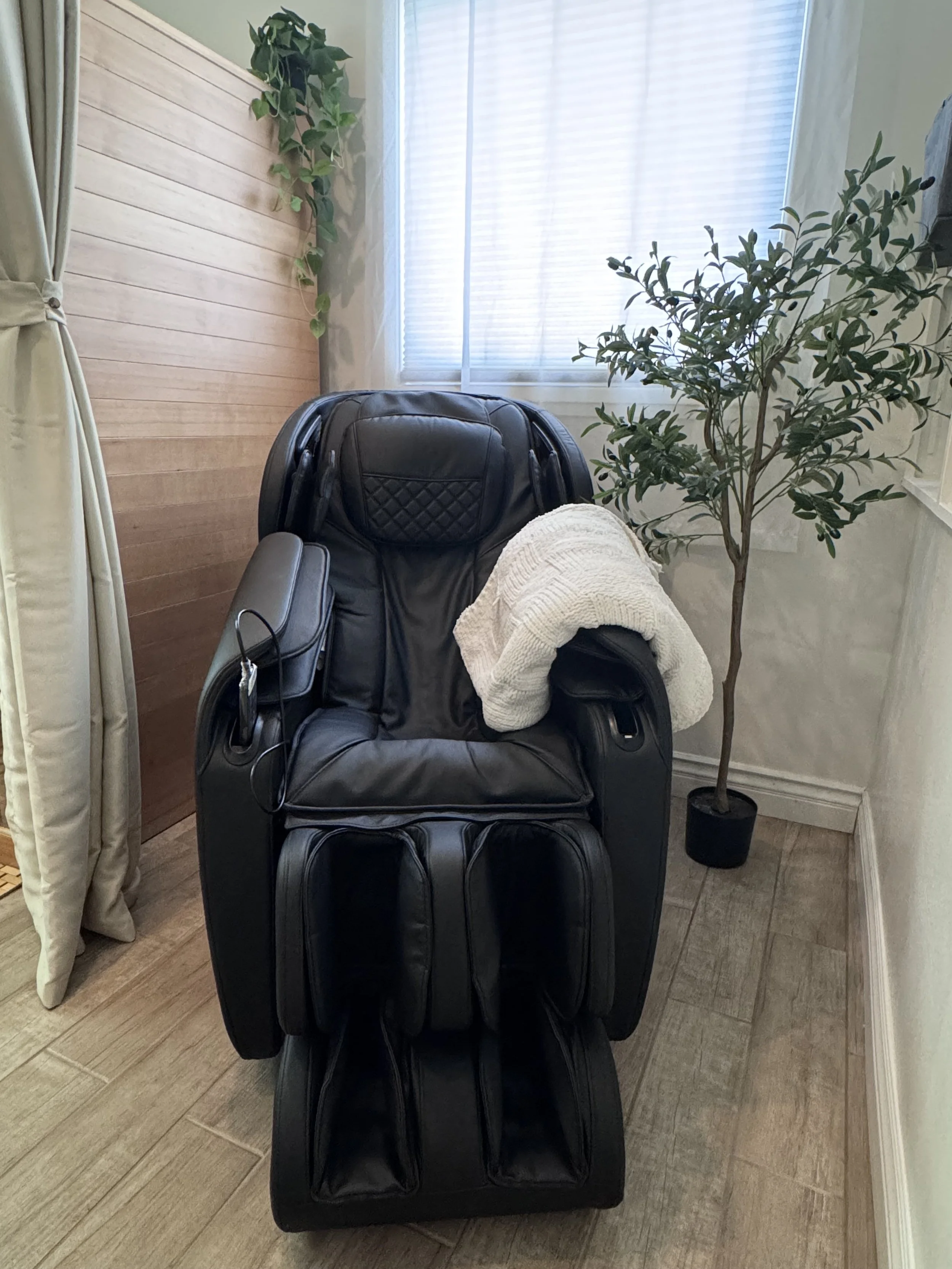 A black zero gravity massage chair in a room with a window, surrounded by green plants and beige curtains.
