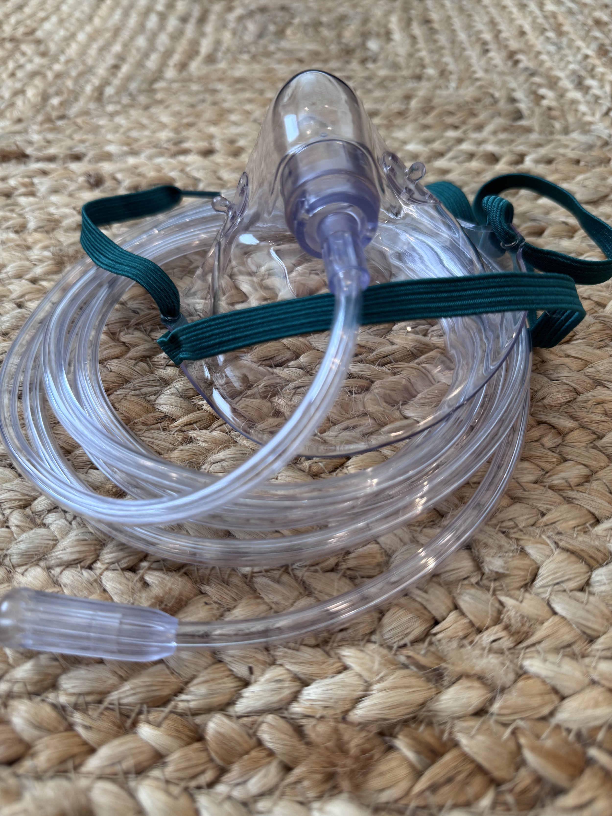 A transparent plastic oxygen mask with a blue elastic strap and a clear tube, resting on a woven beige surface.