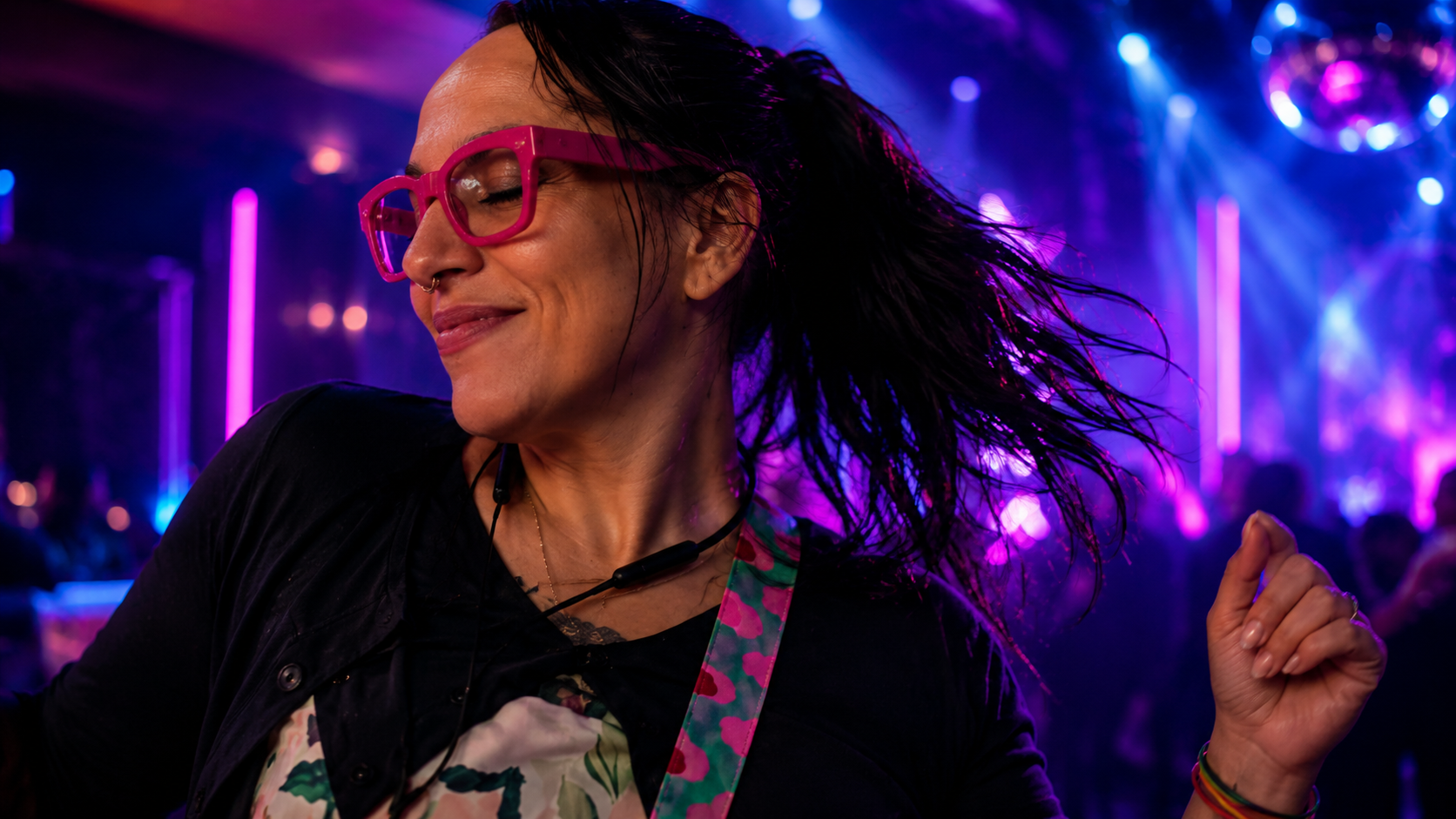 A woman with black hair, pink glasses, and a septum piercing dancing in a nightclub with colorful purple, pink, and blue lights.