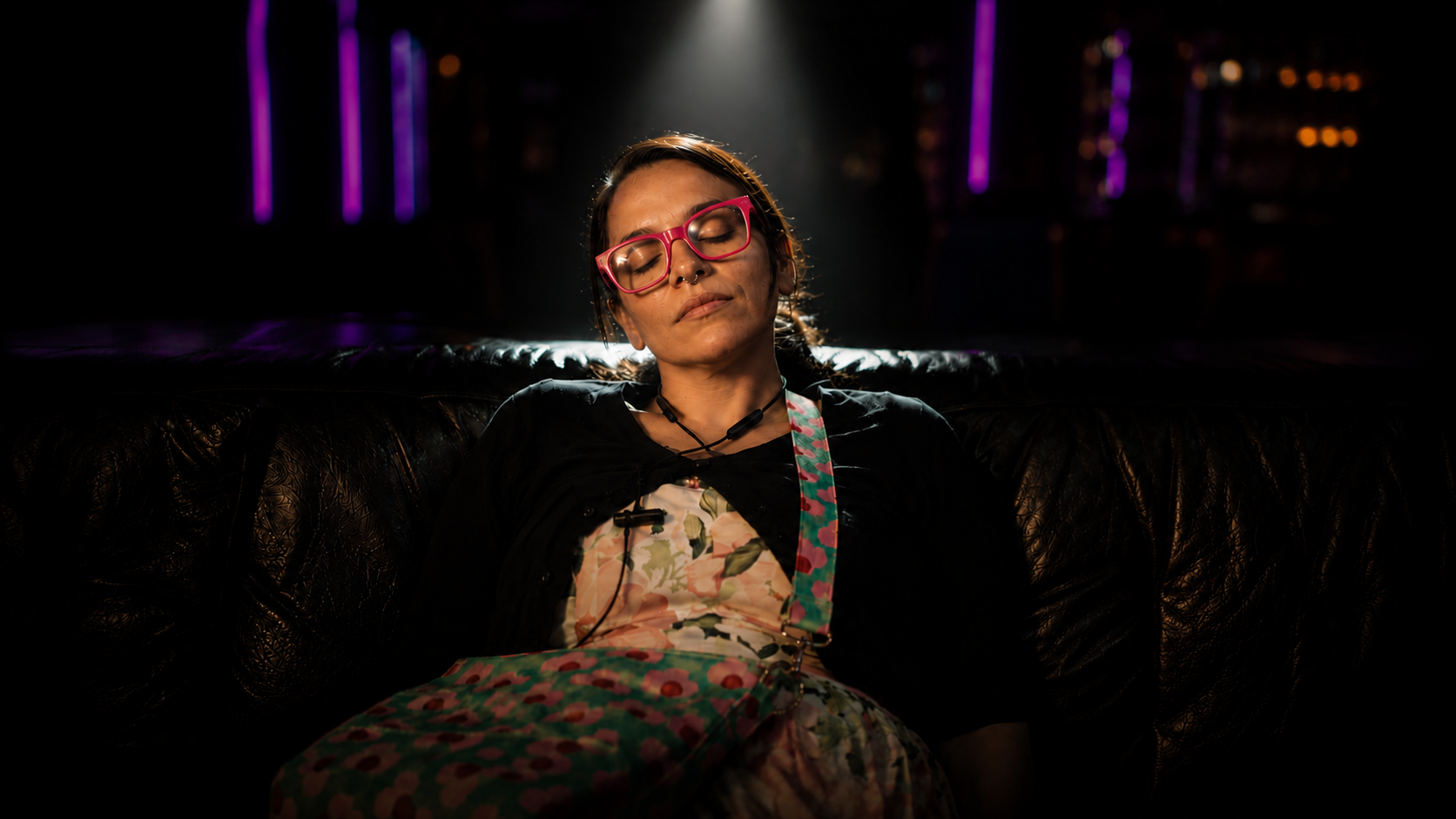 A woman with pink glasses and colorful clothing sitting on a black leather couch in a dark room illuminated by purple lights in the background.