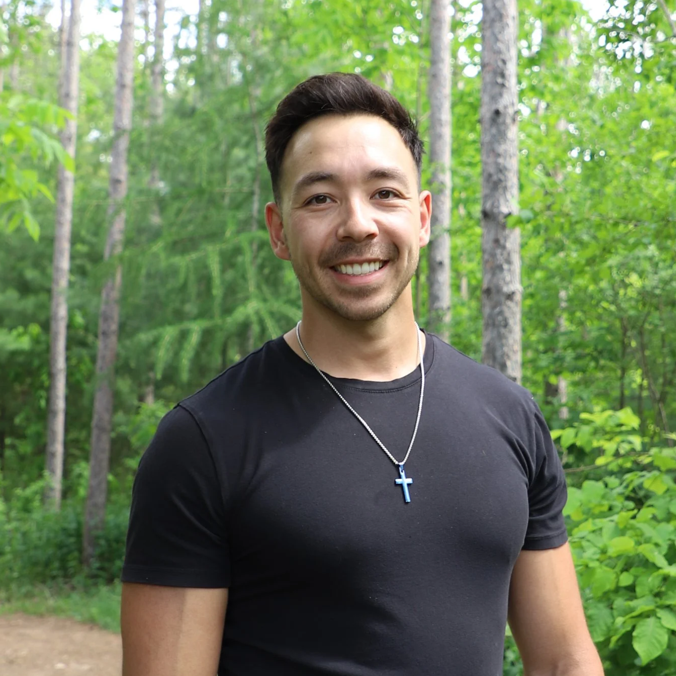 A smiling man with short dark hair wearing a black t-shirt and a silver necklace with a blue cross pendant, standing in a green forest.
