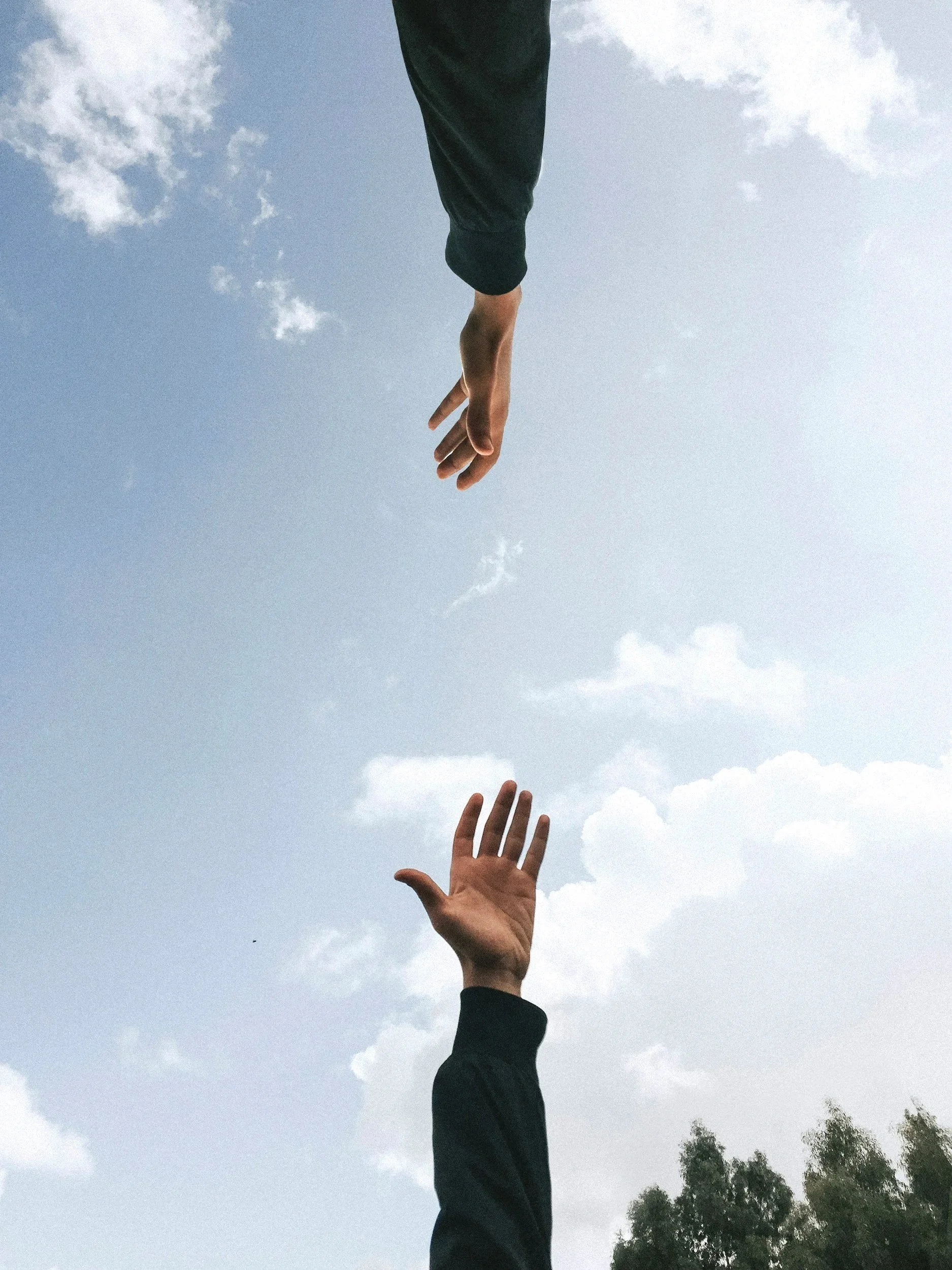 A person extending their hand upward towards the sky, with a blue sky and scattered clouds in the background.