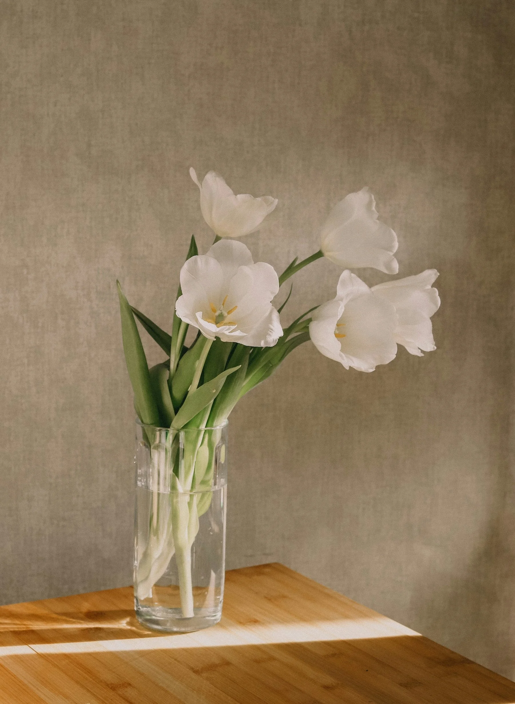 A clear glass vase with white tulips on a wooden surface against a plain wall.