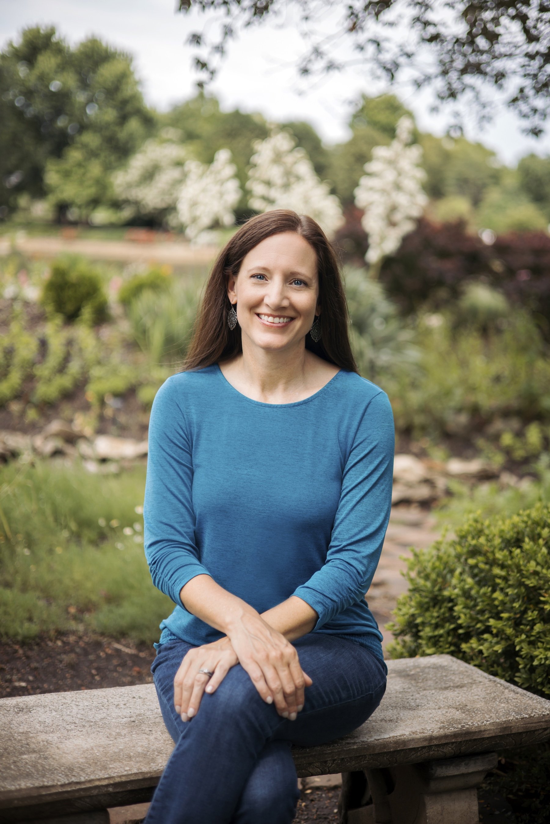 Brooke Nelson, wearing a blue long-sleeve shirt and earrings, sitting on a stone bench in a garden with trees and flowering bushes in the background.