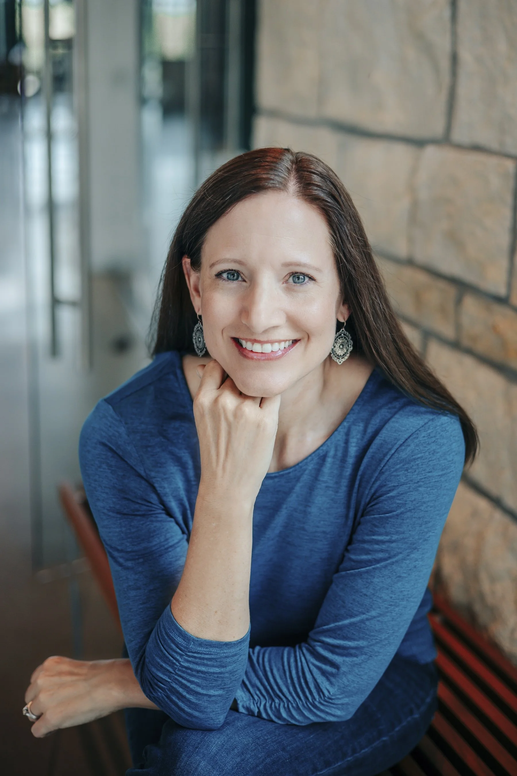 A woman with long dark hair smiling, wearing a blue long-sleeve shirt and large earrings, sitting next to a brick wall.