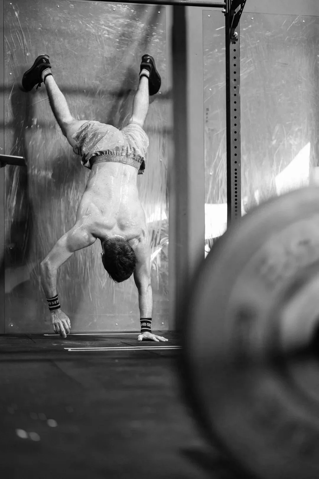 A shirtless man doing a handstand against a wall in a gym, with a barbell on the floor and a weight peg in the foreground.