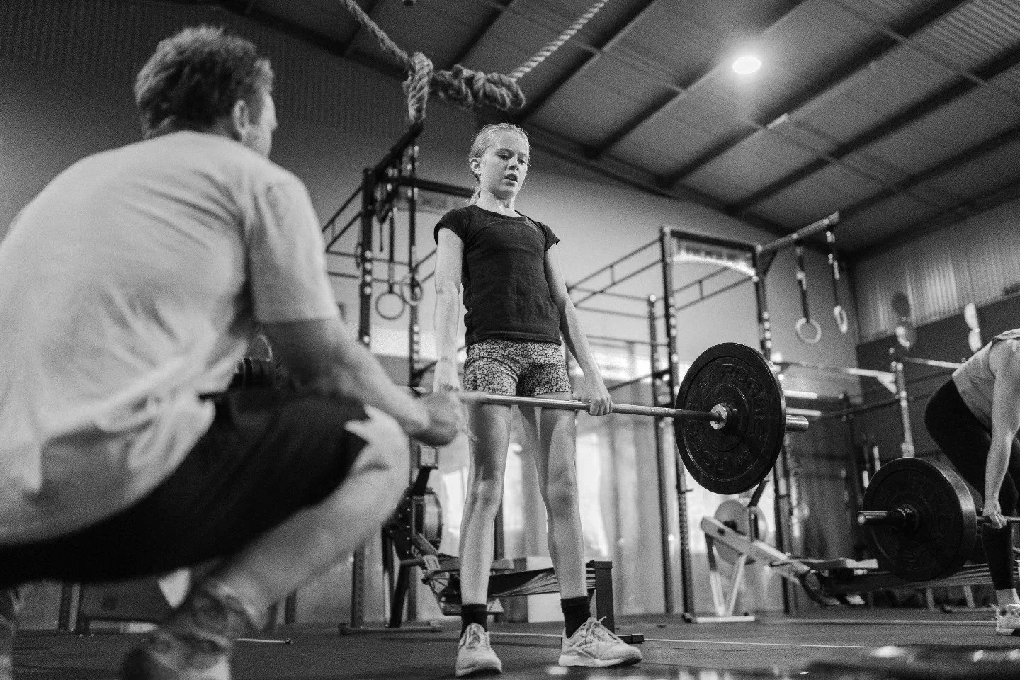 A young girl lifting a barbell with the assistance of a coach or trainer in a gym, with gym equipment and rings in the background.